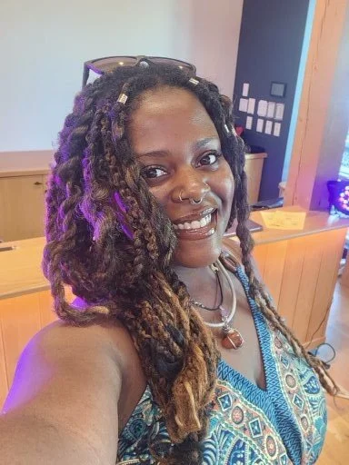 Woman with dreadlocks, glasses on head, piercings, smiling, wearing a colorful patterned top, in a room with wooden furniture and colorful lighting.