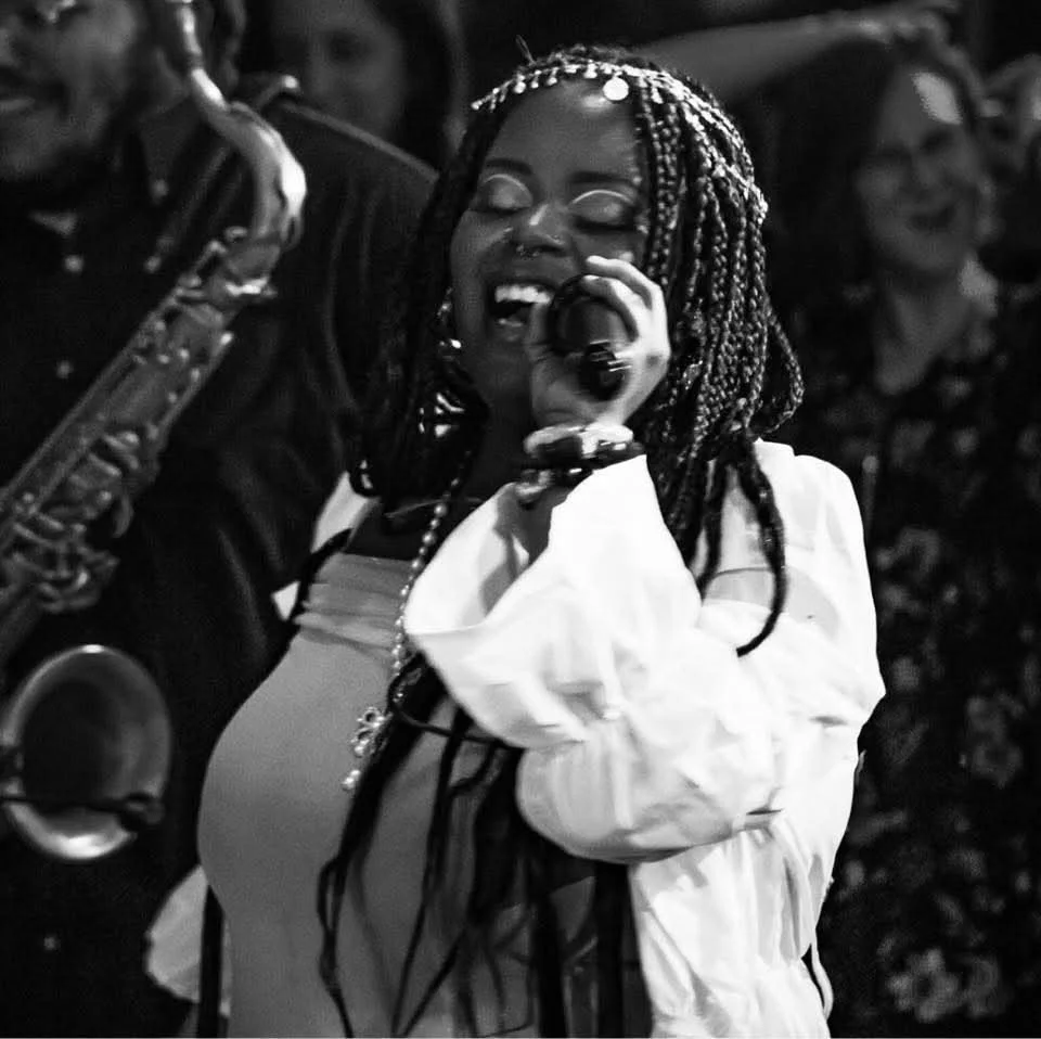 A woman singing into a microphone during a live performance, with a saxophone player beside her and an applause audience in background.