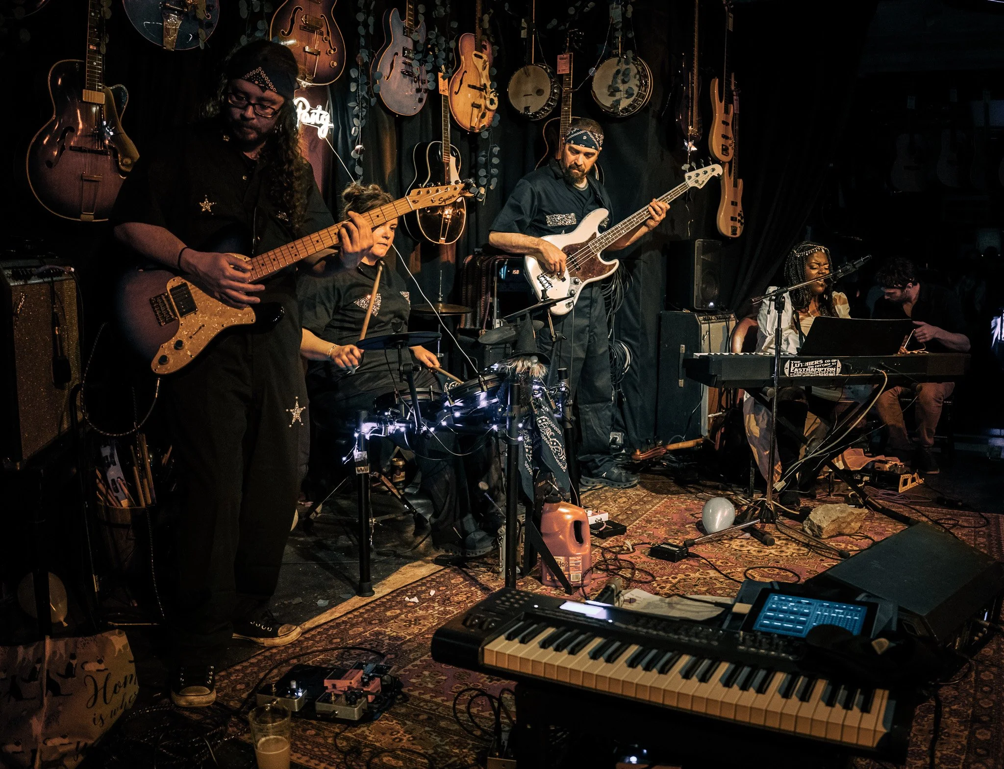 The image shows a band performing on stage with various guitars hanging on the dark wall behind them. There are five musicians visible: a guitarist with long hair and glasses, a drummer, a bassist, a female keyboardist, and a male singer or guitarist