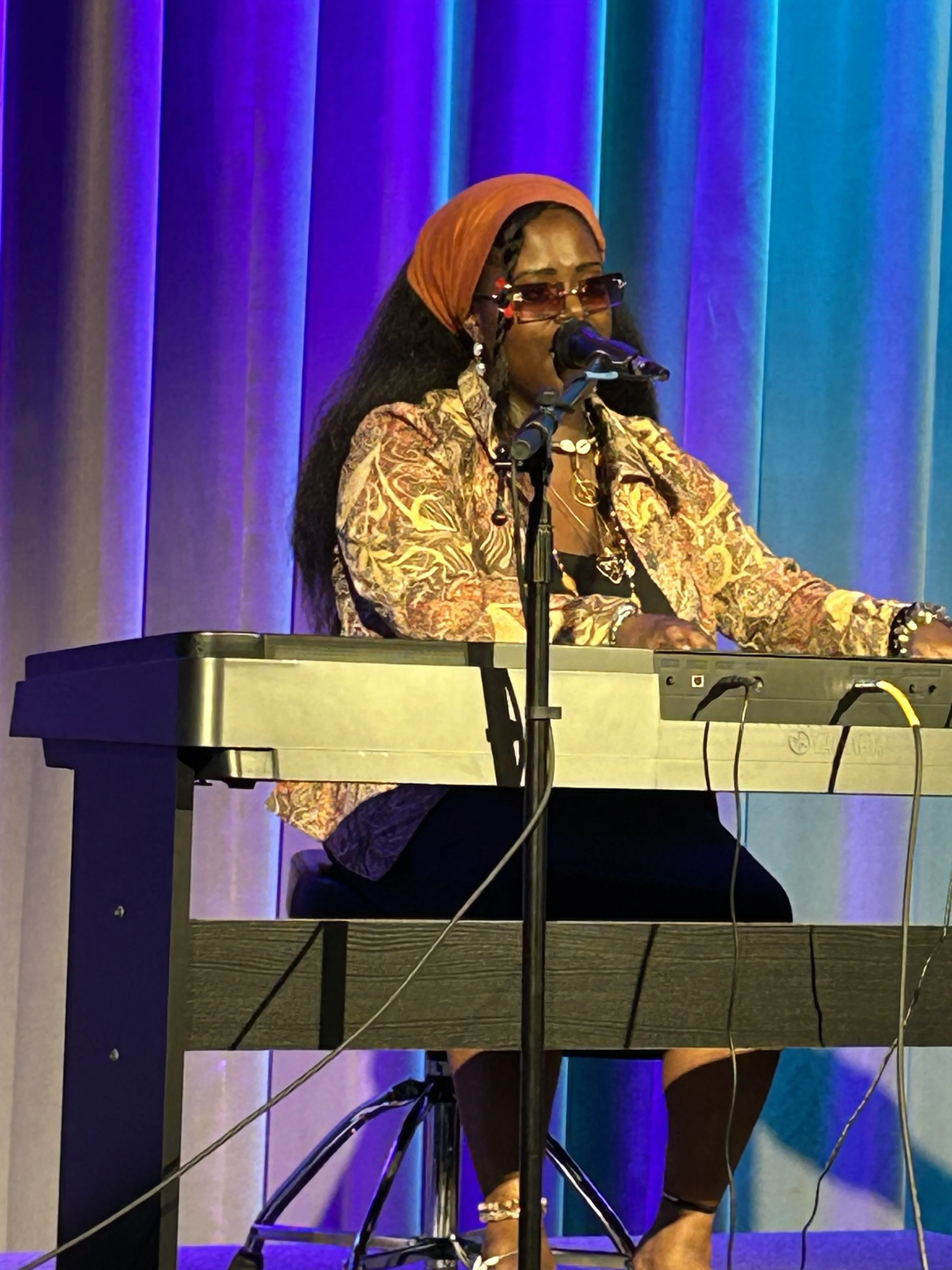 A woman playing a keyboard and singing into a microphone on stage, with a colorful curtain in the background.