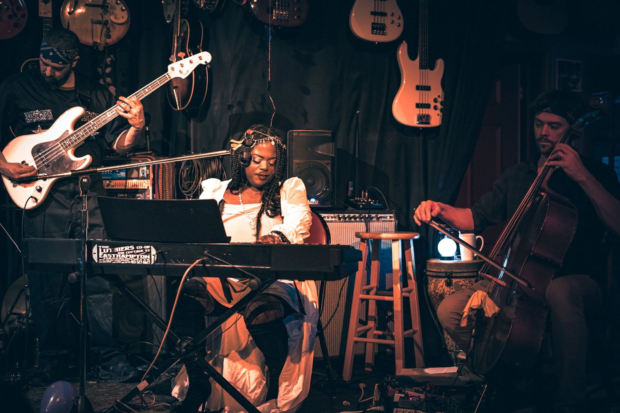 A musical performance on stage with four musicians: a man playing bass guitar, a woman sitting at a keyboard, and two men playing string instruments, in a venue decorated with hanging guitars and basses.