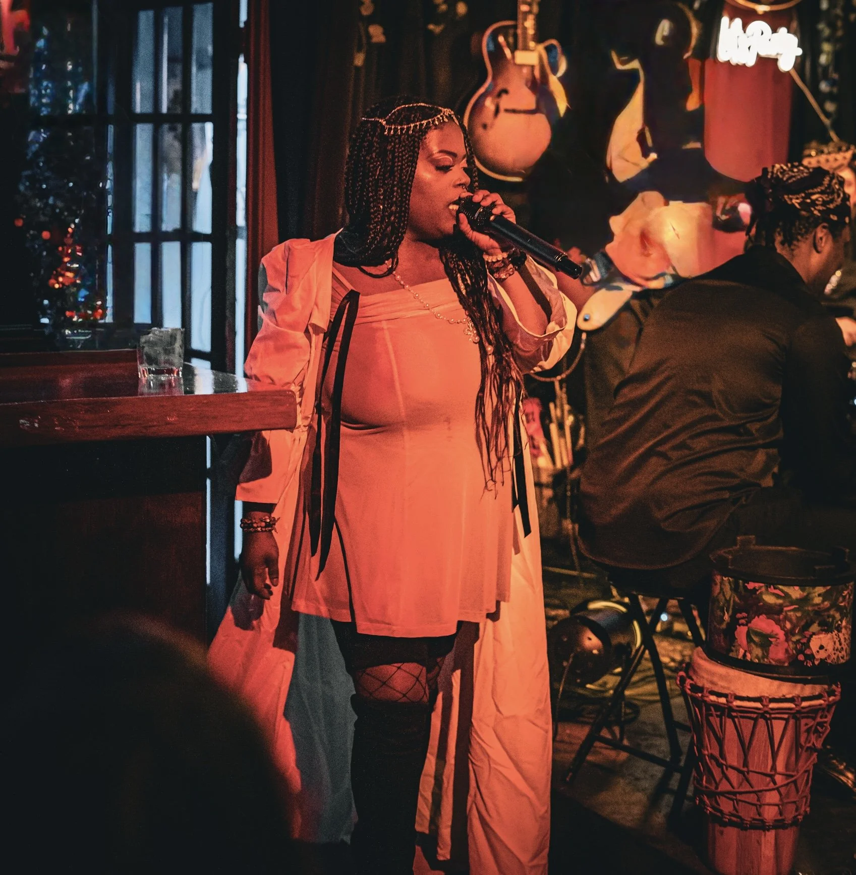 A woman with braided hair is singing into a microphone in a dimly lit bar or club, with guitars and artwork on the wall behind her.