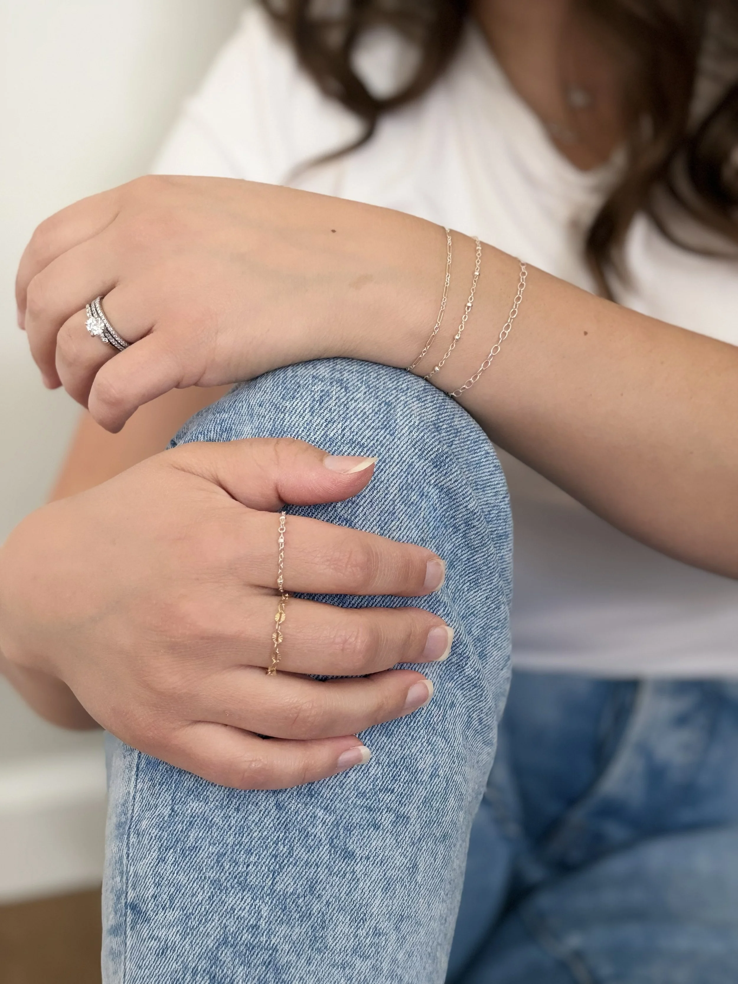Close-up of a woman's hand resting on her knee, wearing a diamond ring, multiple thin chain bracelets, and a gold ring, with her arm and part of her face out of focus in the background.