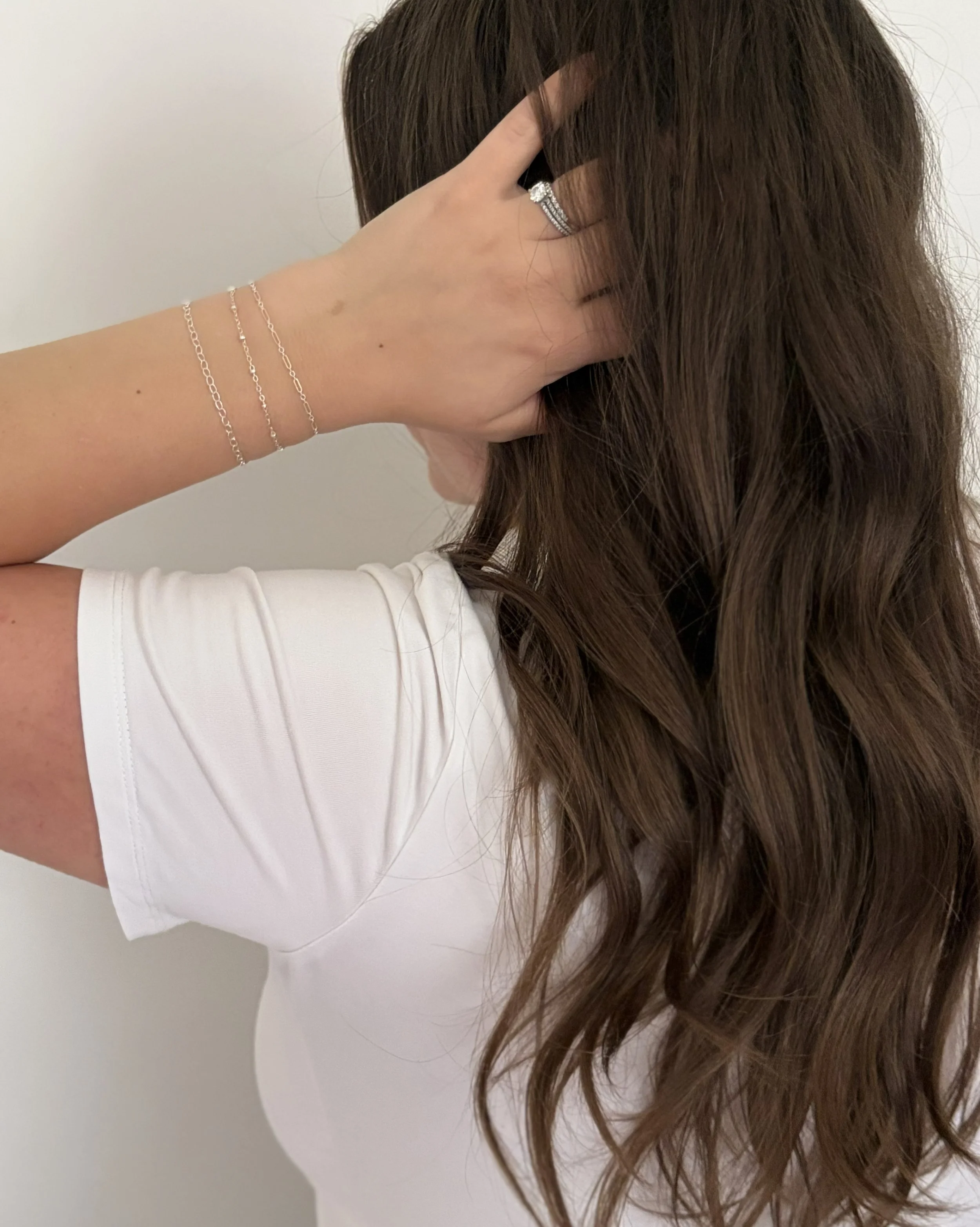 A person's hand with a ring and bracelets resting on their head, with their brown hair covering most of their face. They are wearing a white shirt and are standing against a plain background.