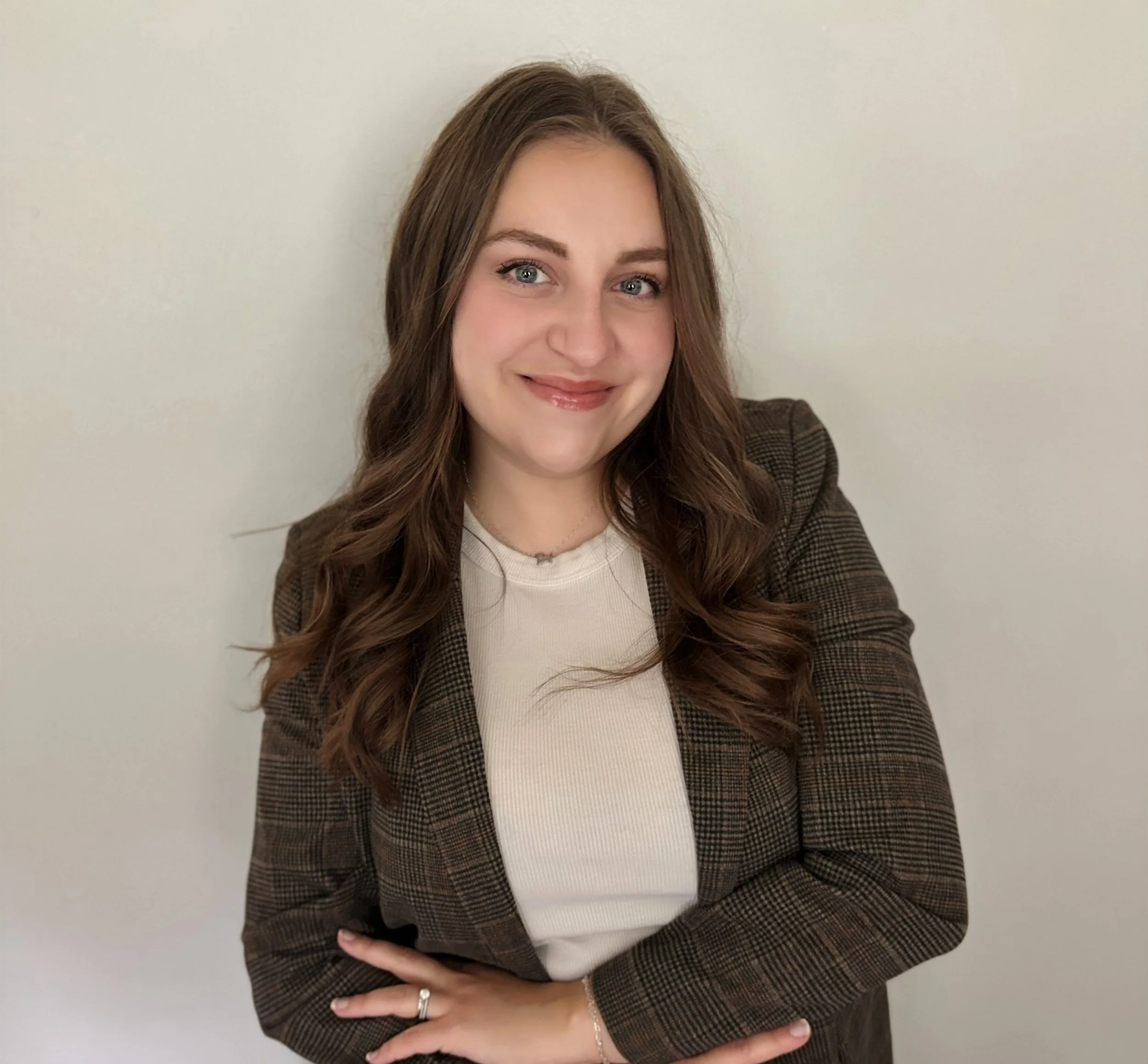 A woman with long wavy brown hair, blue eyes, and light skin smiling, wearing a plaid blazer over a white top, against a plain white wall background.