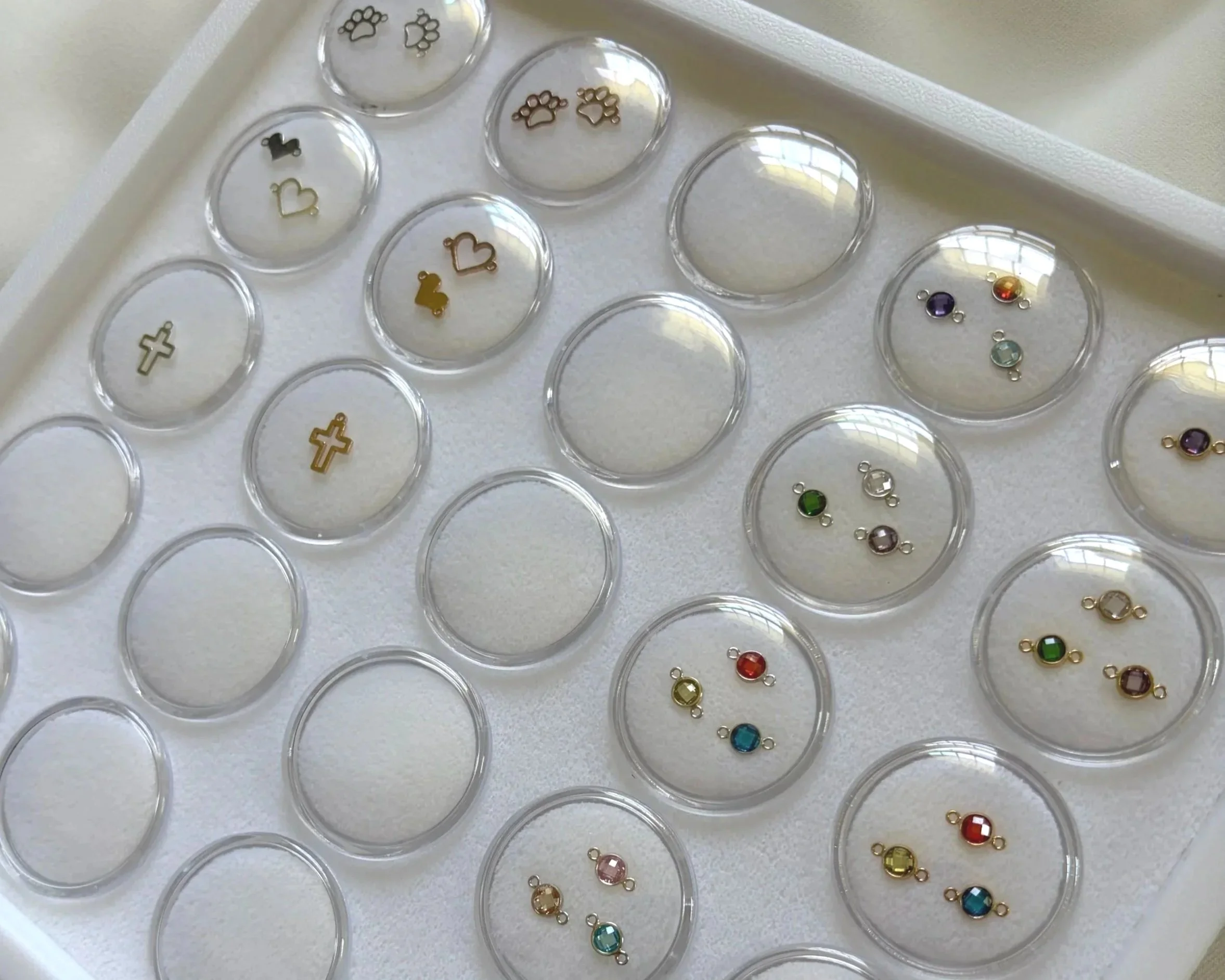 charm and connector case containing small jewelry charms and colorful gemstone charms. Charms include crosses, hearts, and animal paw prints.  Butterfly charm, birthstone, letter initials.