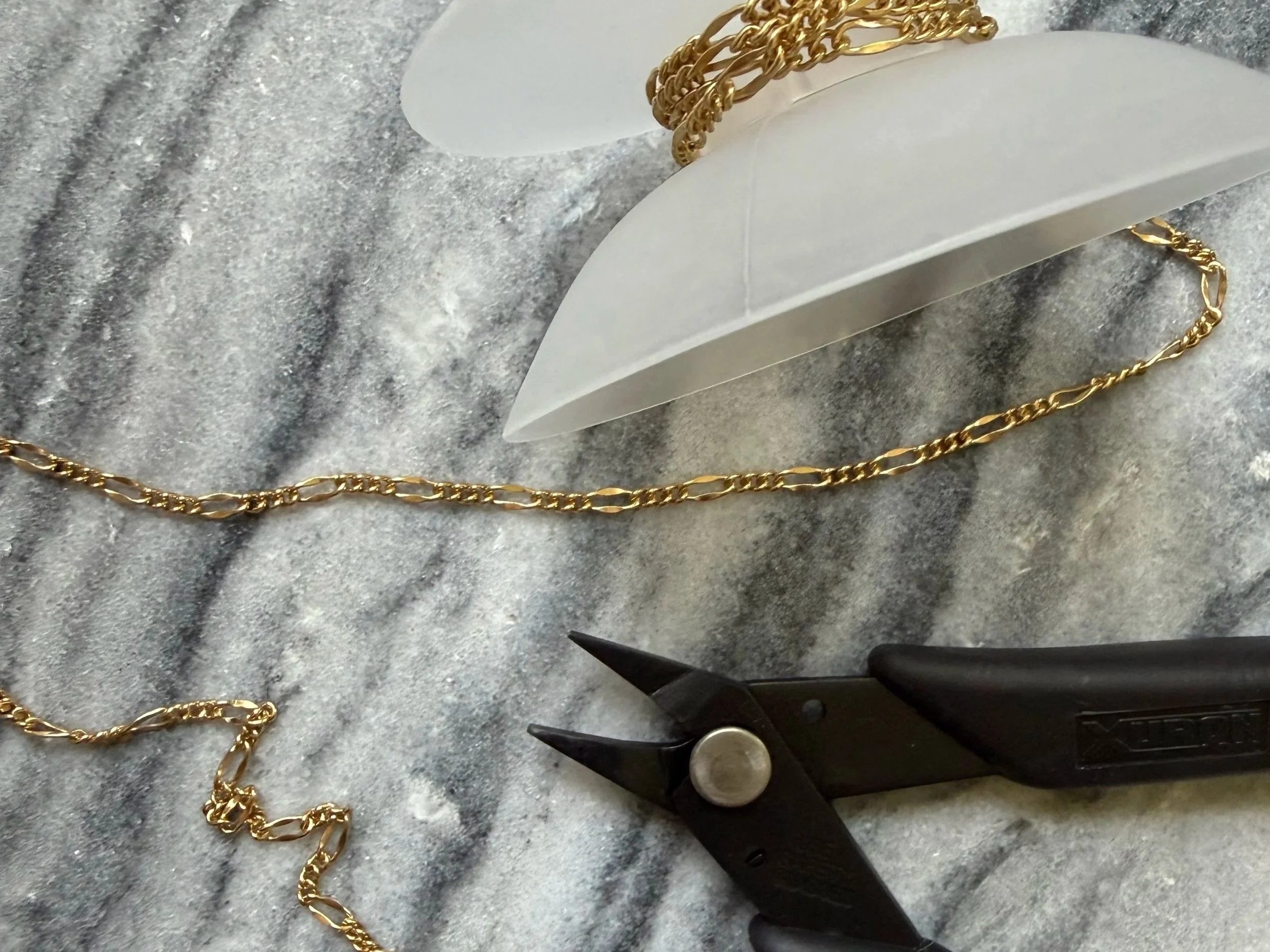Gold chain on a gray marble surface with jewelry-making tools nearby, including pliers and a round object.