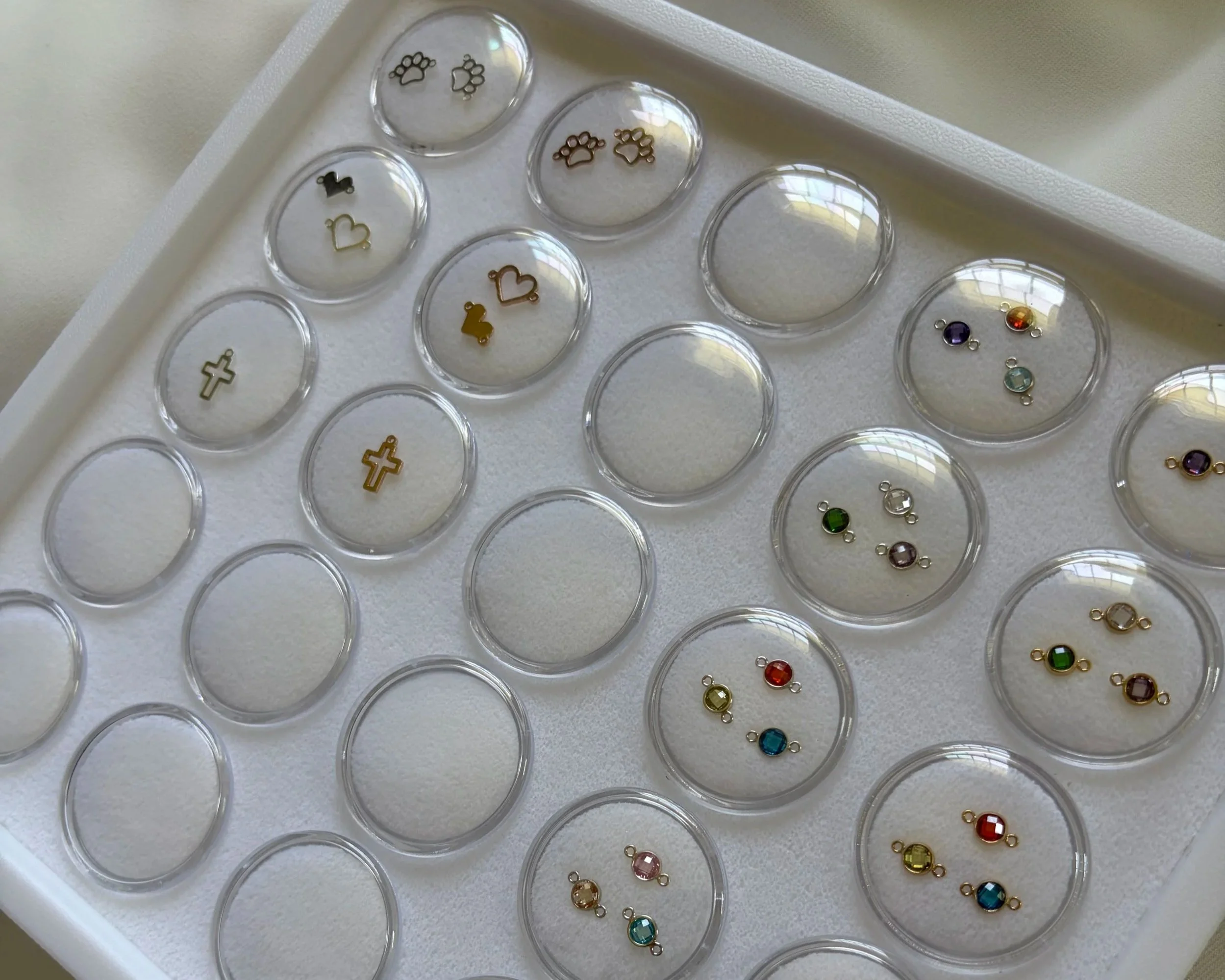 Colorful small charms shaped like hearts, crosses, and paw prints are organized in clear circular containers on a white tray.