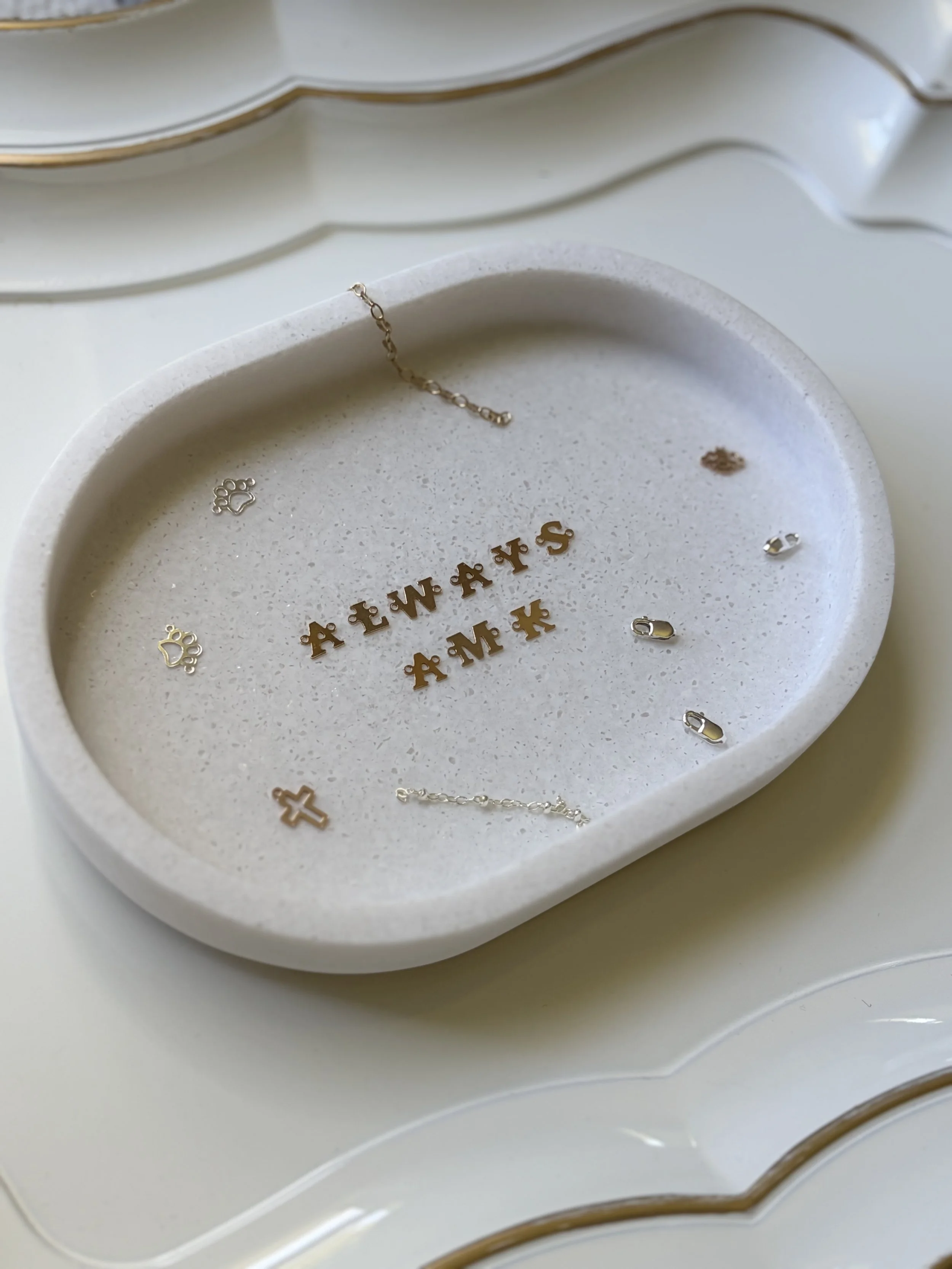 A white ceramic jewelry box with gold accents, containing various small jewelry pieces such as chains, a cross, and alphabet charms.