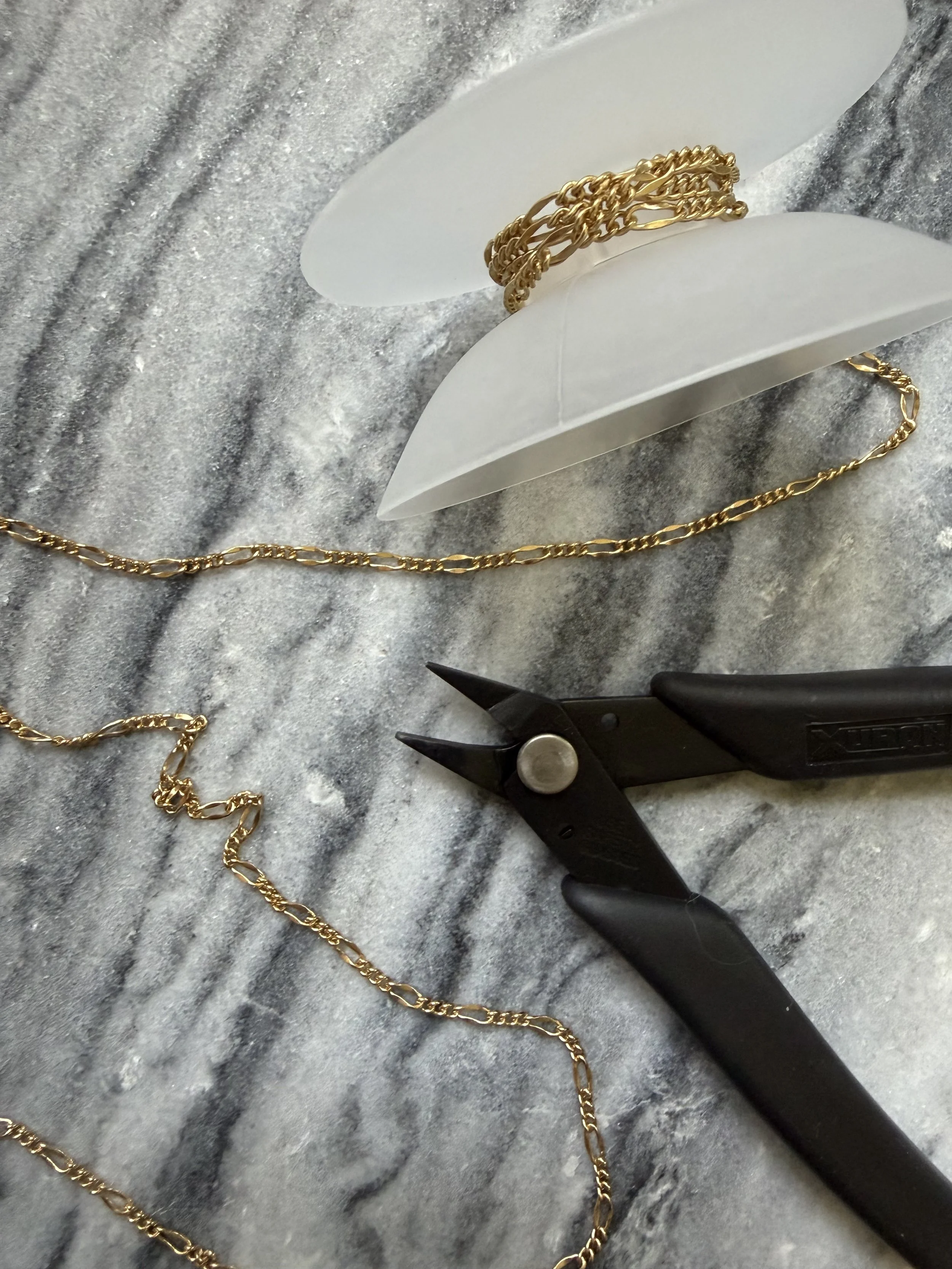 Gold chain necklace, jewelry pliers, and a white plastic jewelry display stand on a gray textured surface.