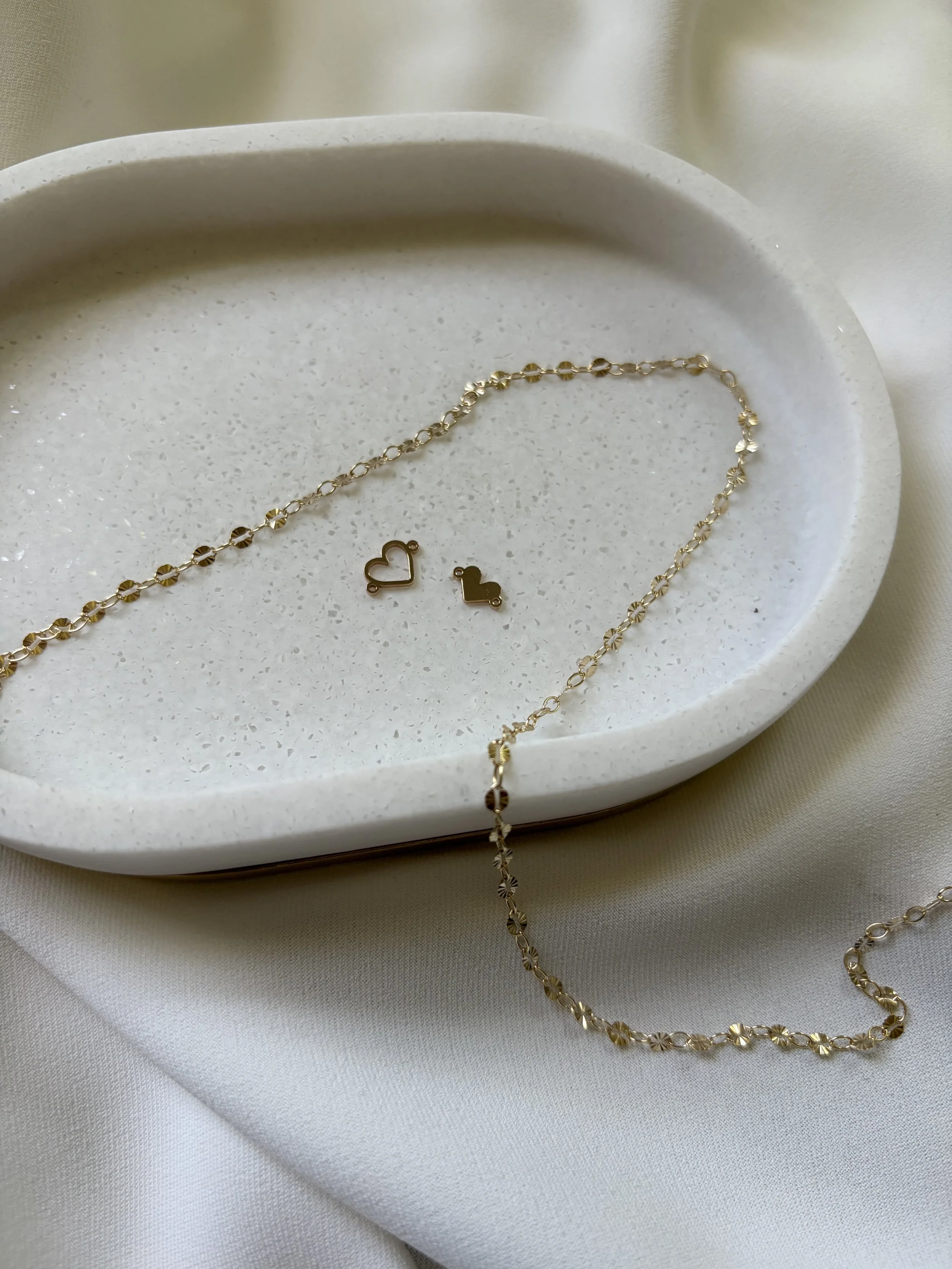 Gold necklace with circular links and heart-shaped and other small pendants laid on a white ceramic dish.