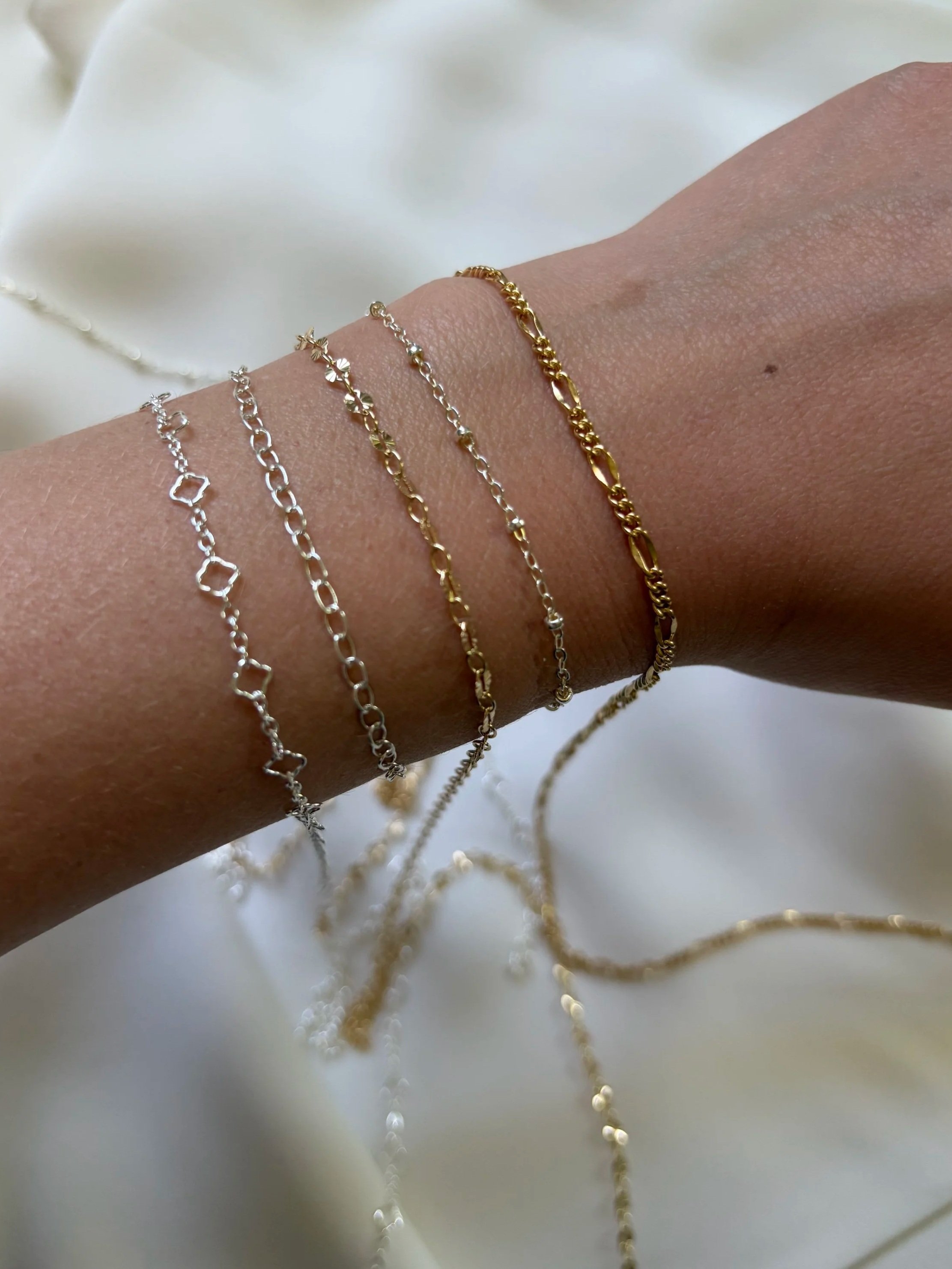 Close-up of a person's wrist displaying multiple delicate chain bracelets in silver and gold tones, set against a soft, white background.
