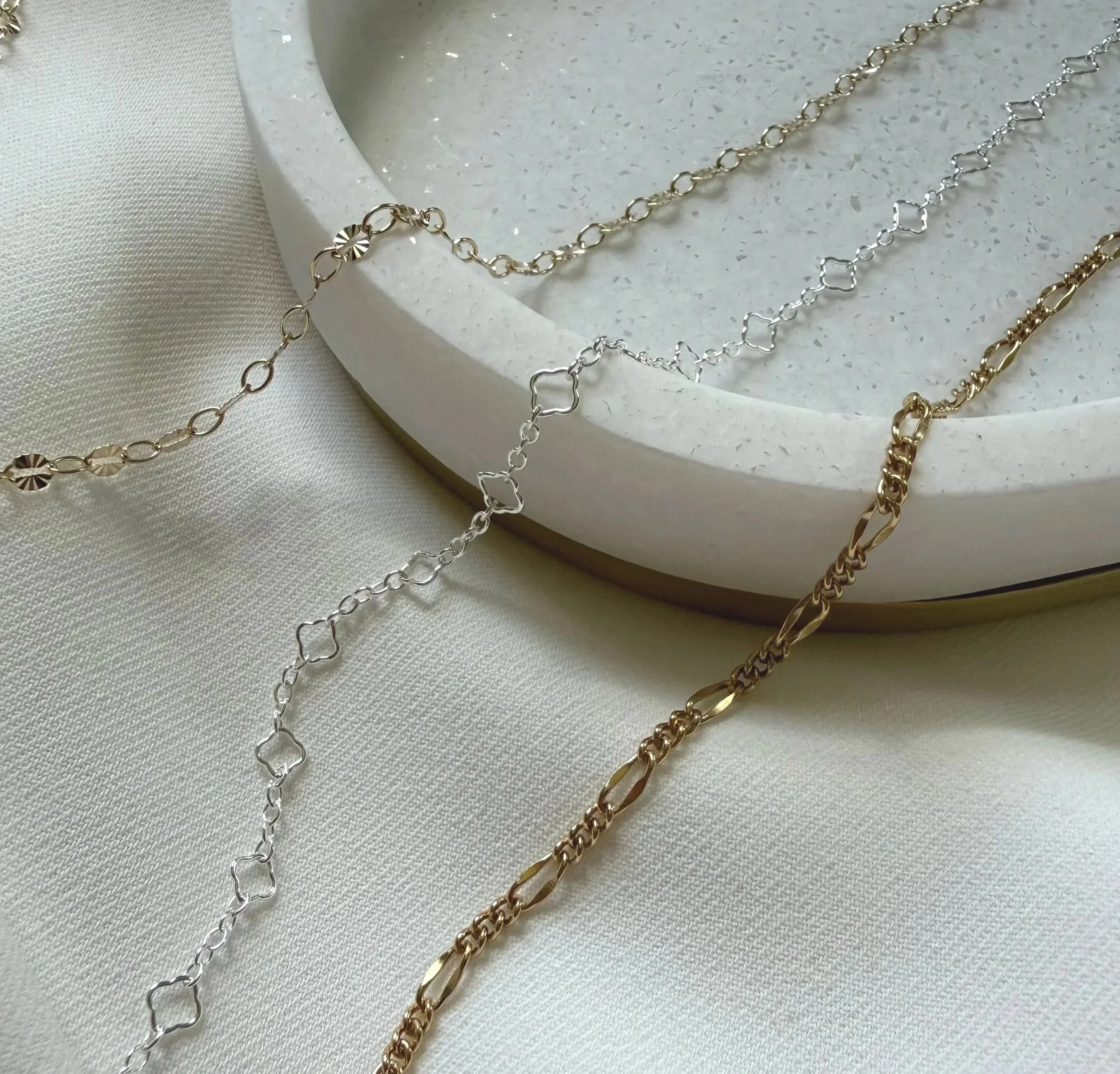 Silver and gold chain necklaces with various link styles laid on a white textured surface and a round white pottery dish.
