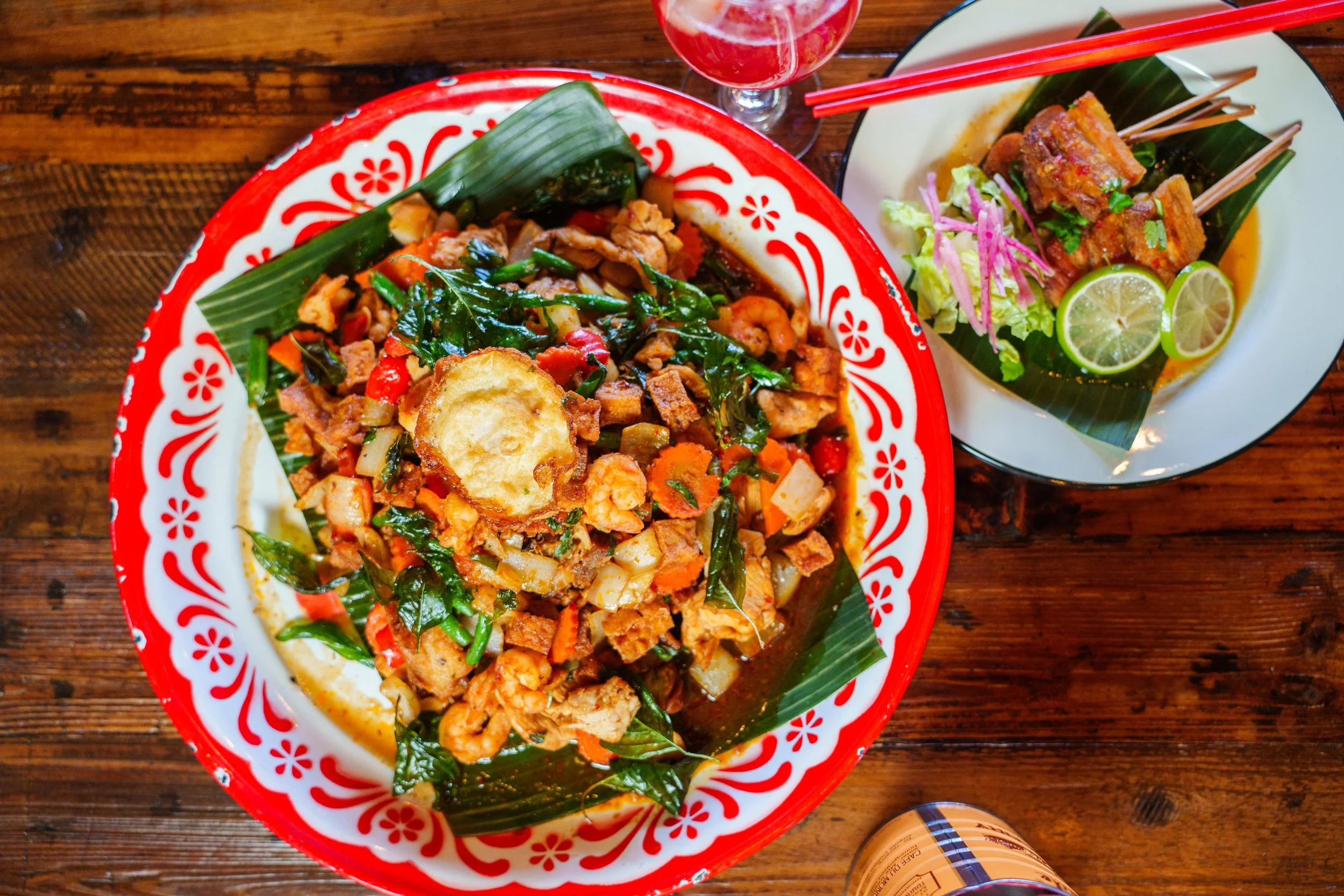 Close-up of two plates of Asian cuisine on a wooden table, one with a colorful noodle stir-fry and the other with skewered grilled meat and lime slices, accompanied by chopsticks and a glass of red drink.