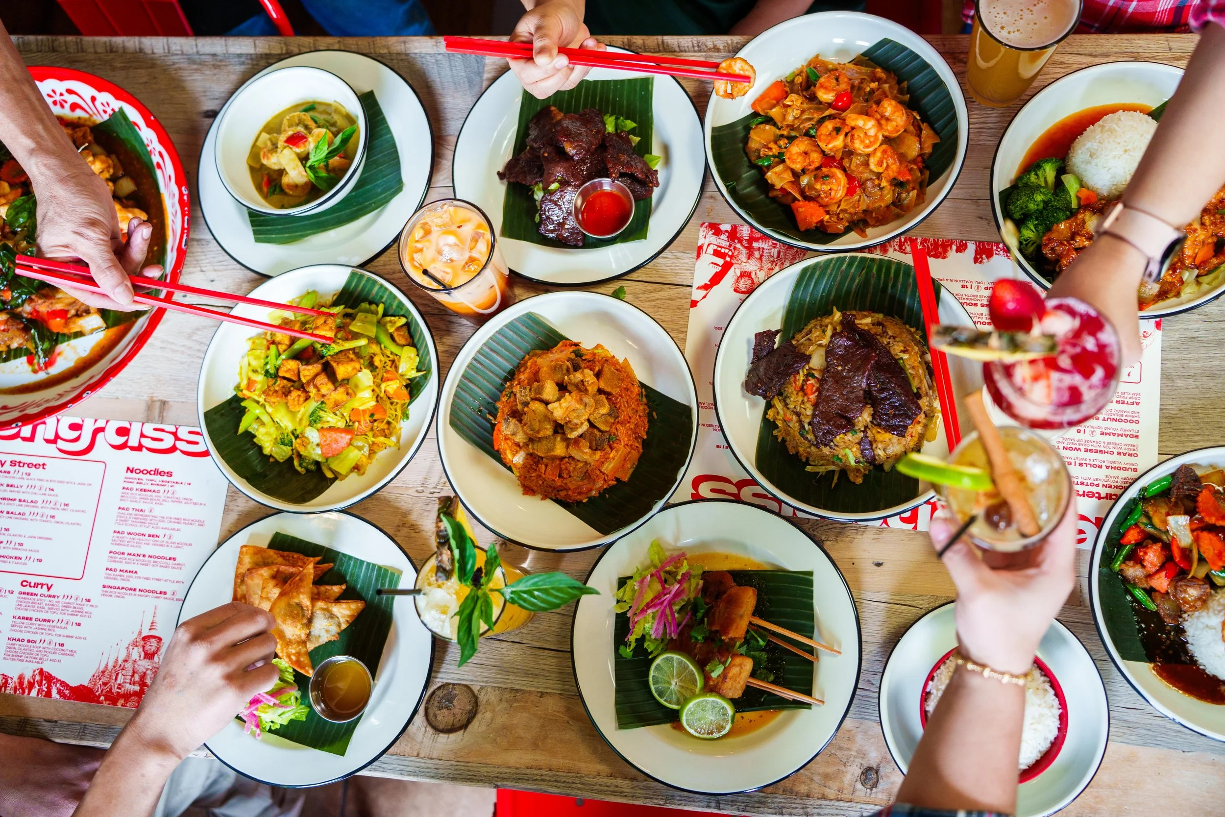A table filled with various Asian dishes, including bowls of rice, shrimp, vegetables, and grilled meats, with people's hands reaching for food and drinks.