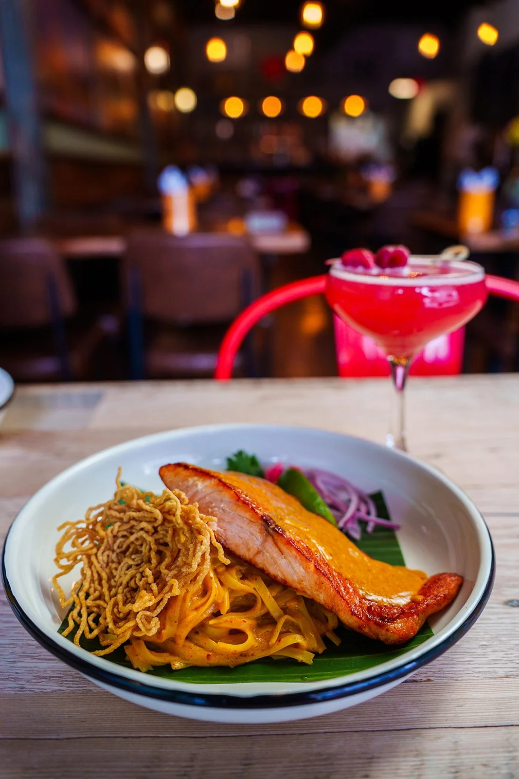 A bowl of Asian noodle dish with grilled chicken, fried crispy noodles, and fresh vegetables, with a pink cocktail garnished with raspberries on a wooden table in a cozy restaurant.