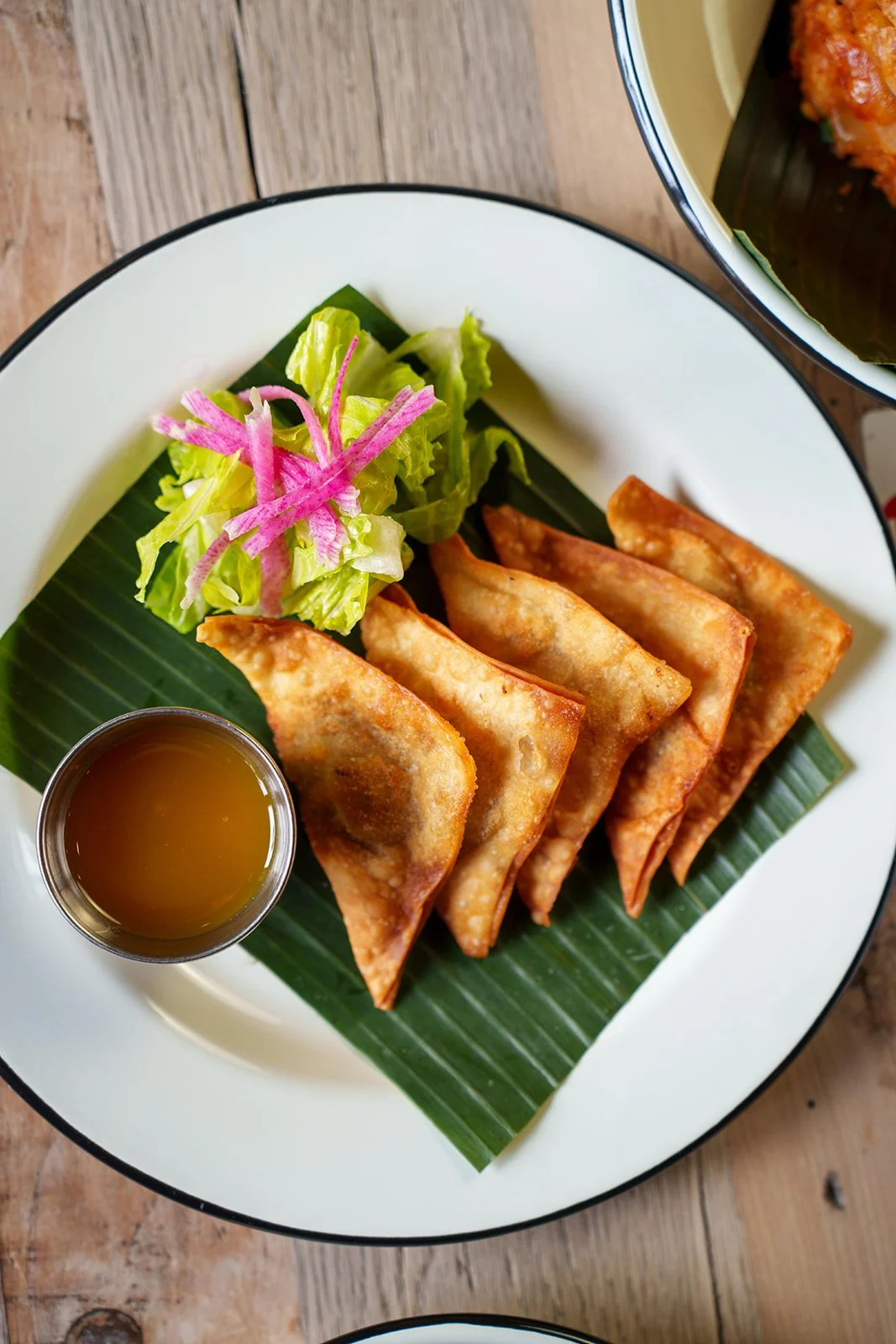 Plate with five fried dumplings, a small cup of dipping sauce, and a side salad of lettuce and pink pickled vegetables on a banana leaf.