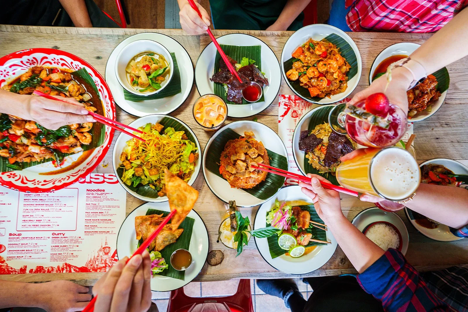 A top-down view of a table with various Thai dishes, drinks, and hands holding chopsticks and beverages during a meal.