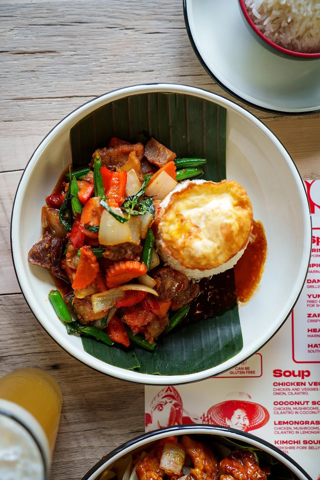 A bowl of stir-fried vegetables with beef, a dollop of rice topped with a baked egg, and a slice of orange sauce on the side. There is a bowl of white rice and a glass of yellow beverage on a wooden table.