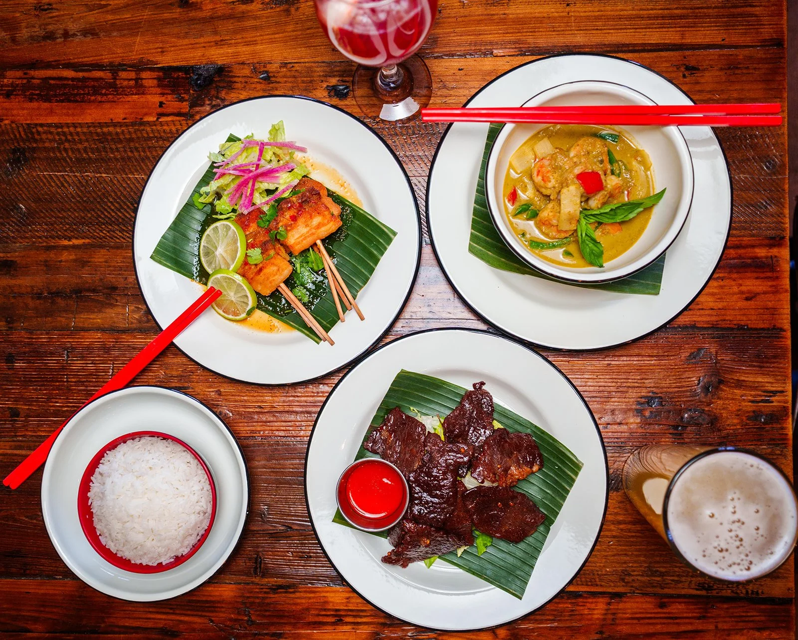 Assorted Asian dishes on a wooden table, including rice, grilled meat, a creamy curry with vegetables and meatballs, and skewered grilled meat, with red chopsticks, a glass of red drink, and a glass of beer.