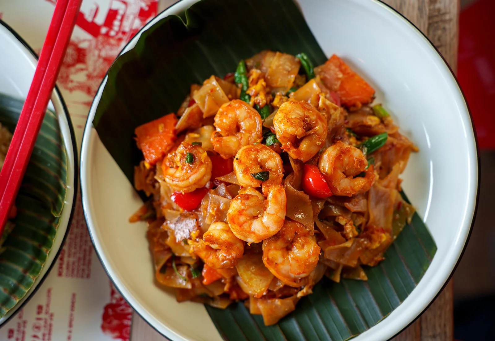 A bowl of shrimp, vegetables, and noodles in a sauce, served in a traditional Asian dish with a banana leaf lining.