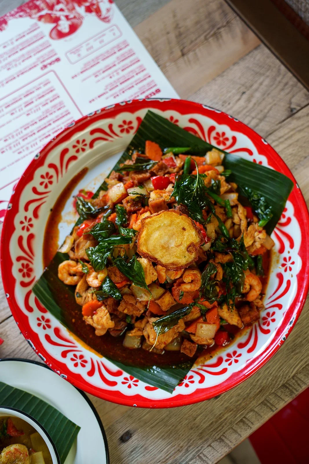 A colorful plate of Asian stir-fry with vegetables, shrimp, and shrimp paste or fried garlic on top, served on a decorative red and white plate with banana leaf accents.