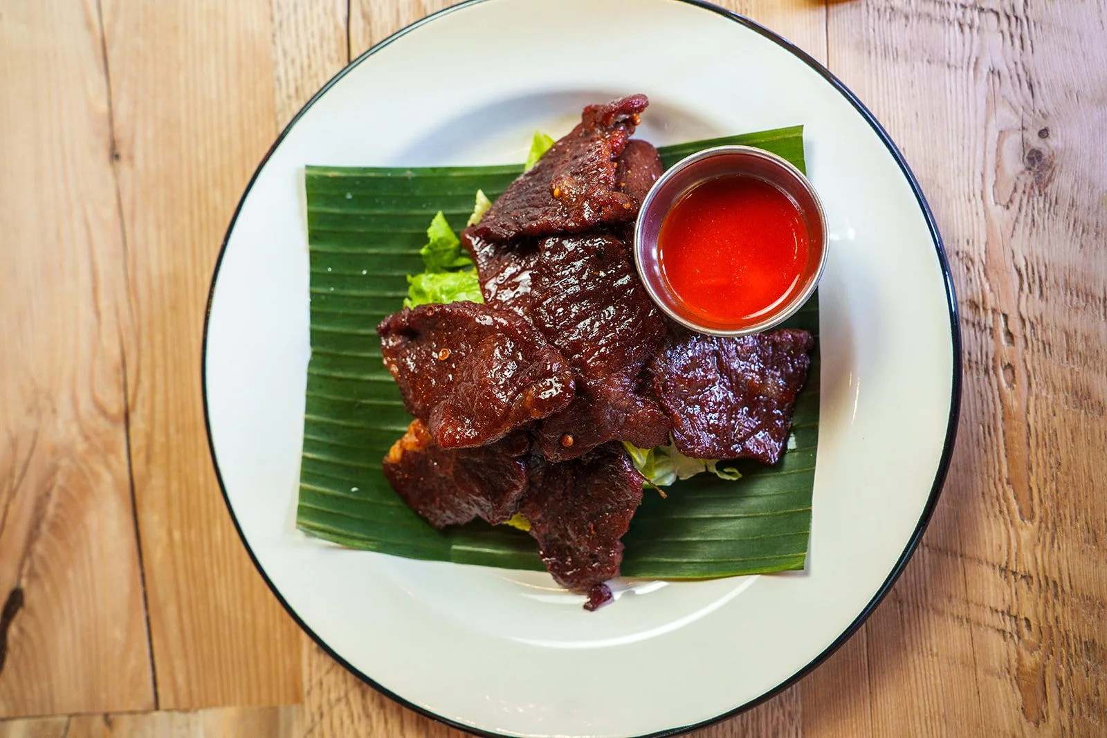 Plate of cooked beef slices on a banana leaf with small cup of red sauce on a white and black-rimmed plate, on a wooden surface.