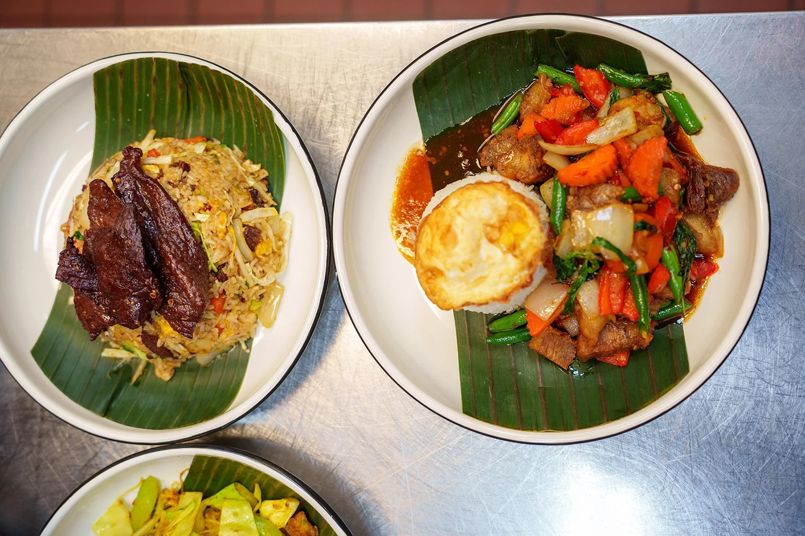 Two bowls of Asian cuisine on a metal surface, one containing fried rice with meat, and the other with stir-fried vegetables and a fried egg on top, served on green banana leaf inserts.