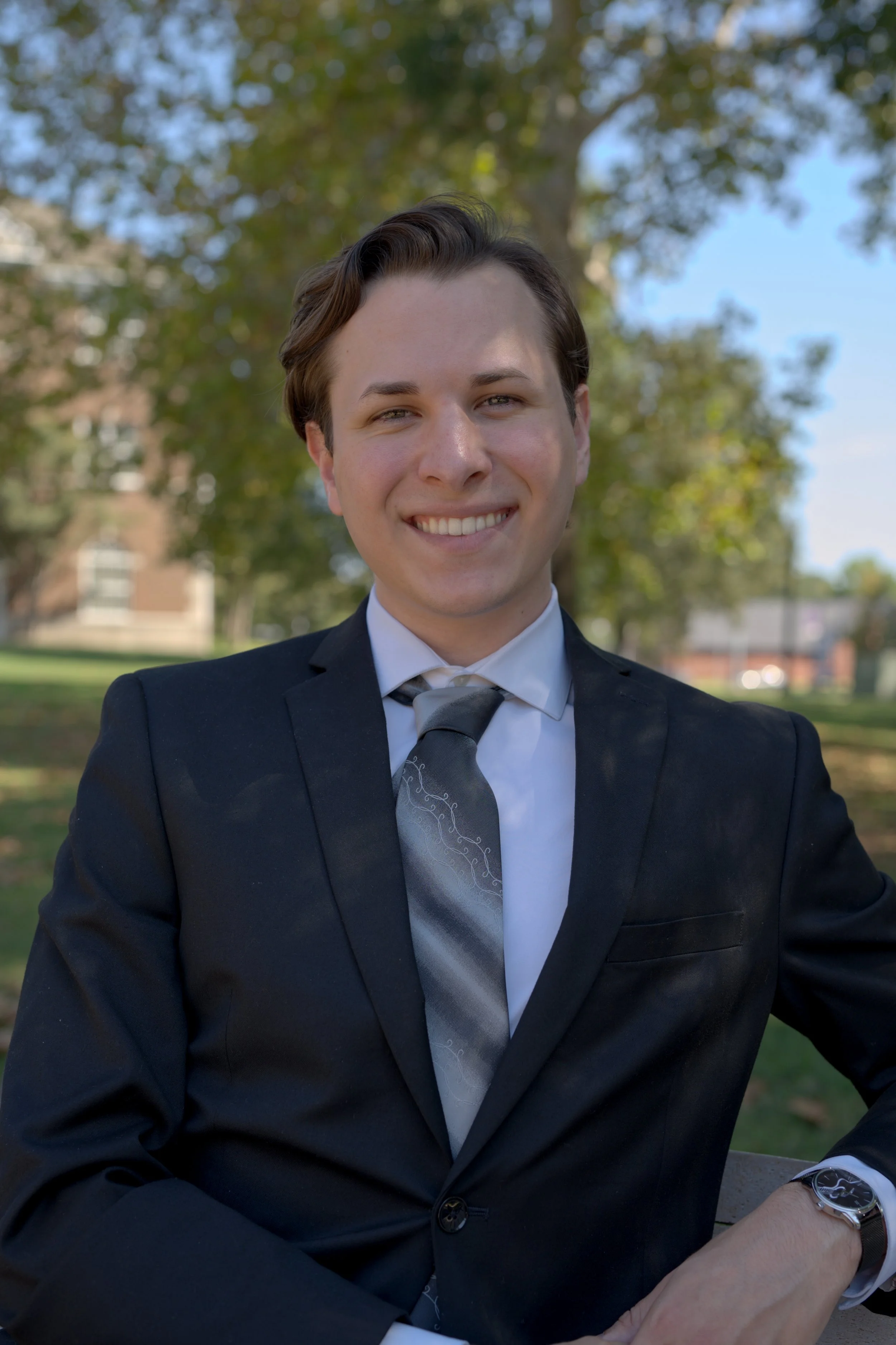 Aidan Kolokithas in a black suit, white shirt, and gray patterned tie sitting outdoors in front of a blurred green and blue background, smiling at the camera. He can help protect your family with valuable insurance policies.