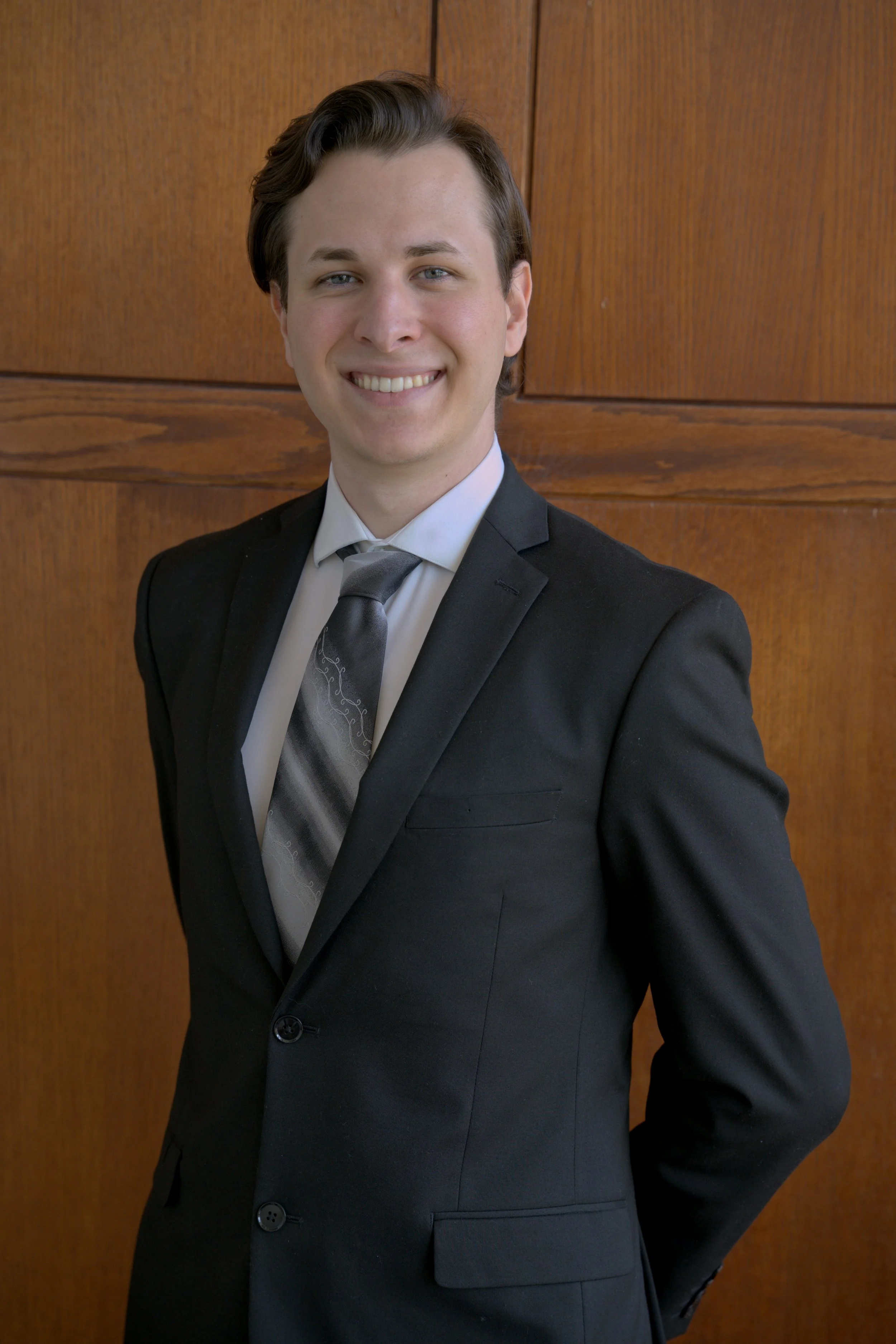 Aidan Kolokithas in a black suit with a patterned tie, standing against a wooden wall, smiling and looking at the camera. He is a licensed Arkansas insurance agent and can help protect you and your family.