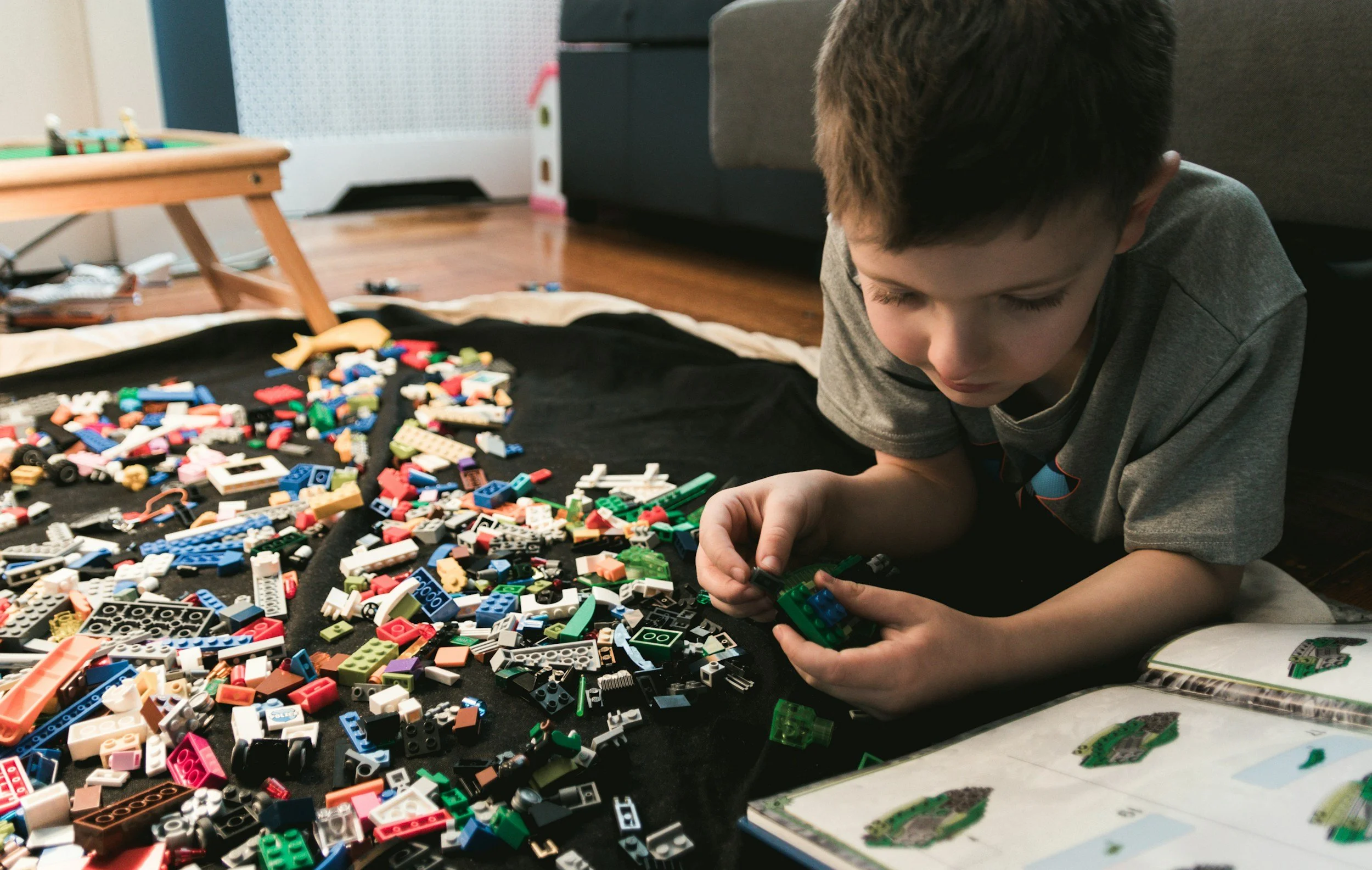 A young boy with short dark hair is laying on the floor, focused on assembling a colorful Lego set. There is a large collection of assorted Lego bricks spread out on a black mat in front of him, with some booklet pages showing Lego model instructions nearby. The room has a wooden floor and a gray sofa is visible in the background.