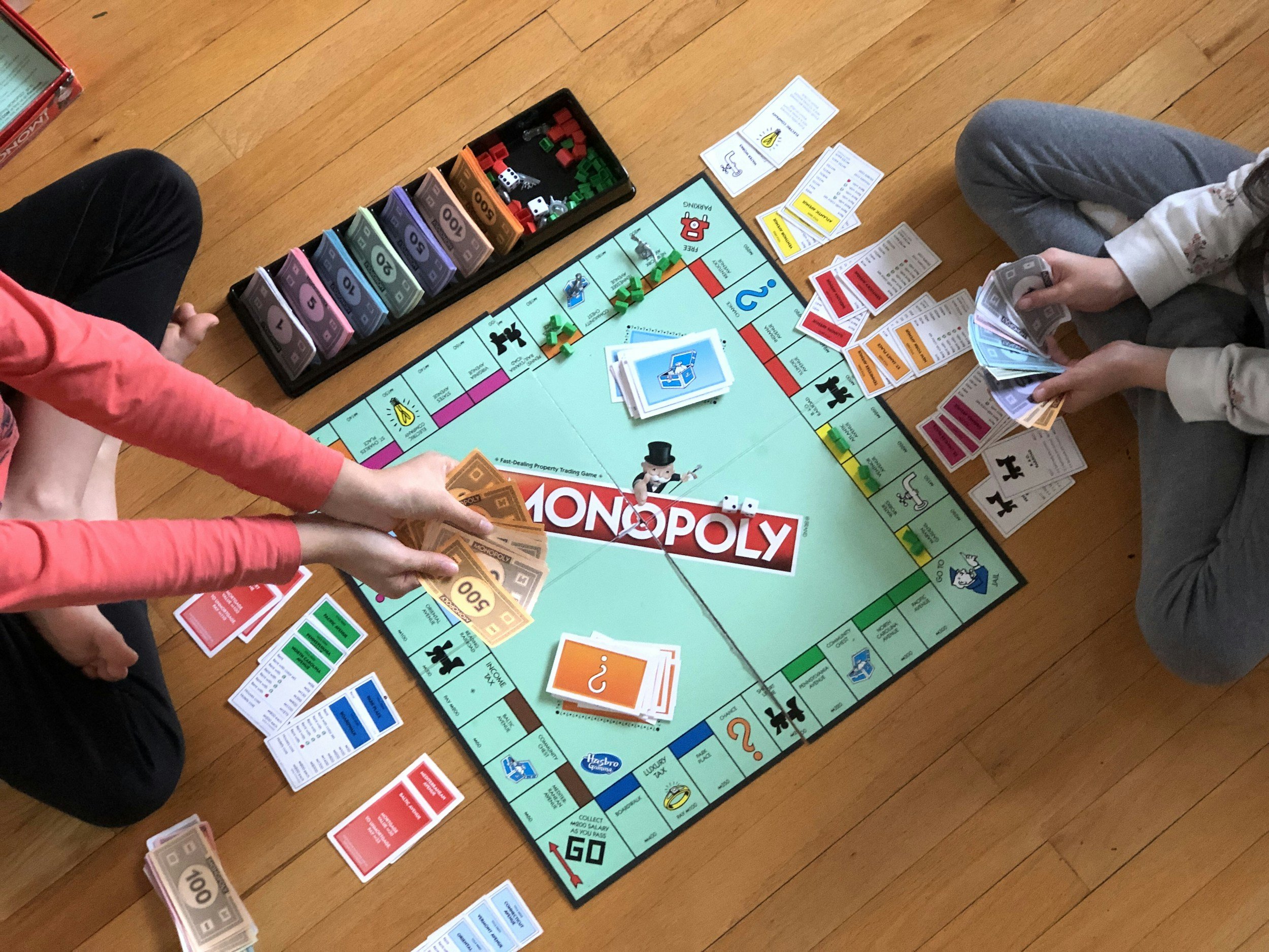 People playing Monopoly with game pieces, money, and property cards on a wooden floor.