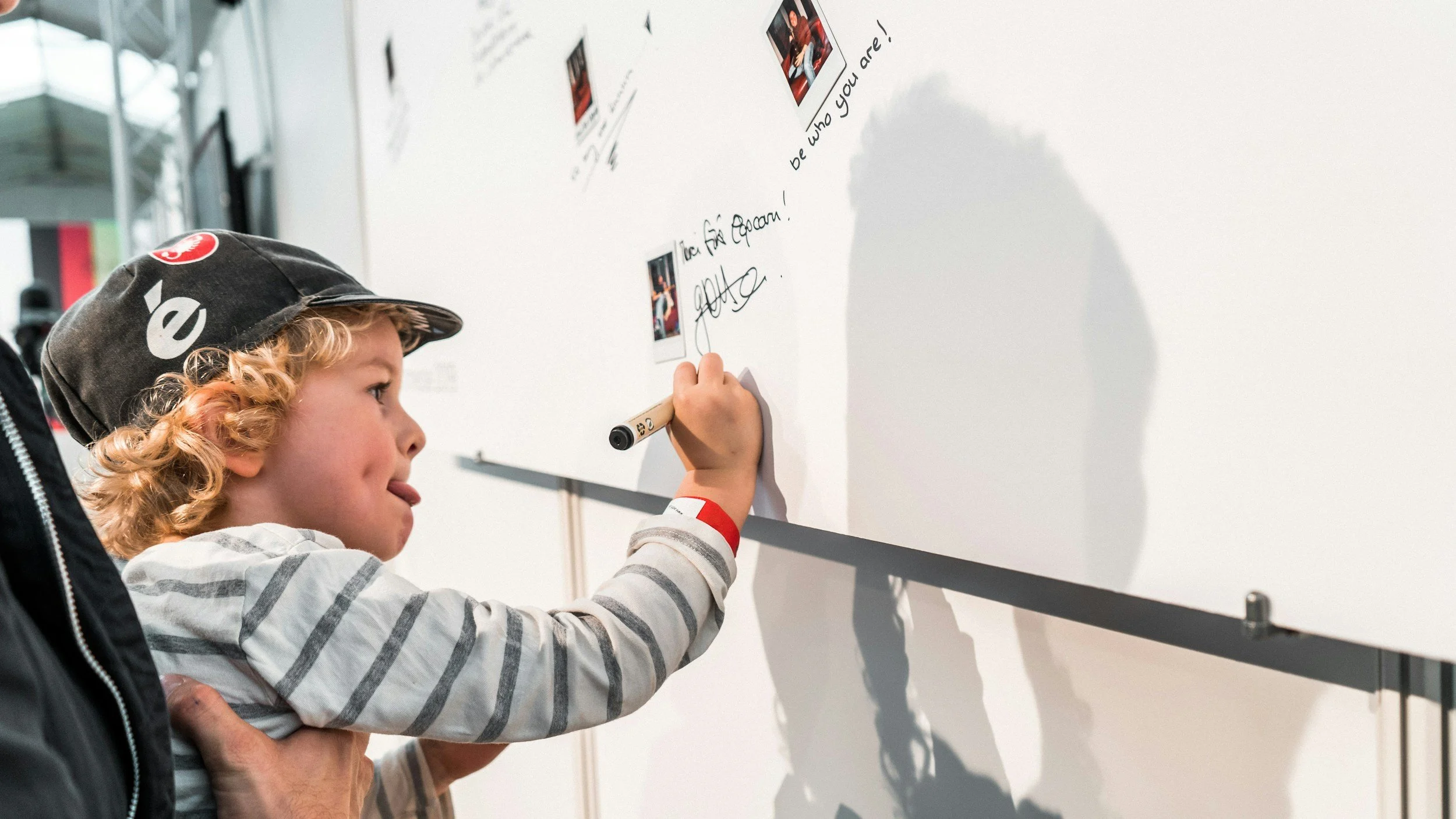 A young boy with curly blonde hair and a cap is writing on a whiteboard with a marker, while an adult's hand supports him. The whiteboard has photos and handwritten notes, and the boy is focused on his writing.