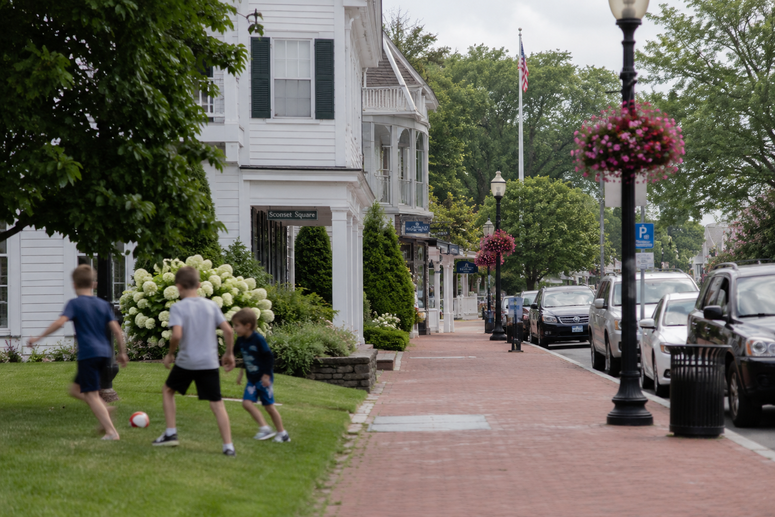 Children playing with a soccer ball on a grassy lawn in a quaint town street lined with shops, flowering hanging baskets, lampposts, and parked cars, with trees and an American flag in the background.