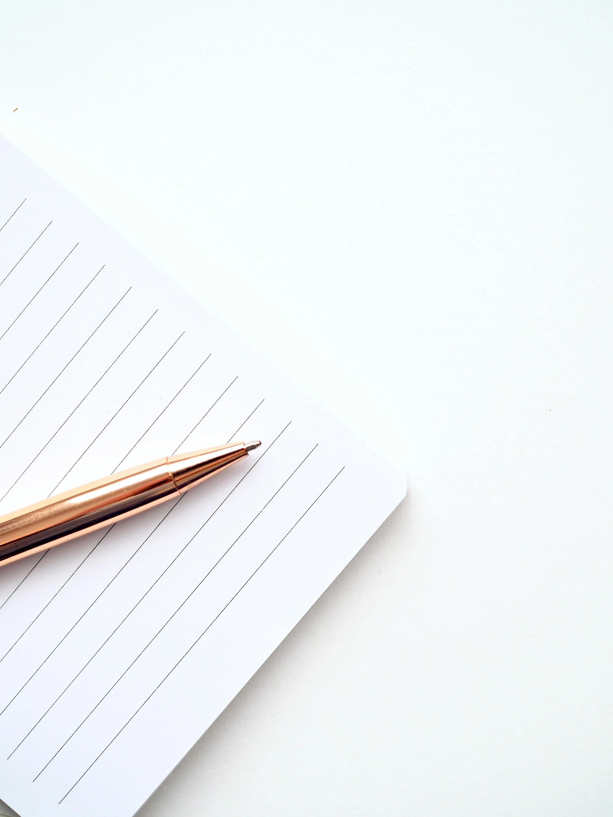 A rose gold pen resting on a white lined notepad with several pages, placed on a white surface.