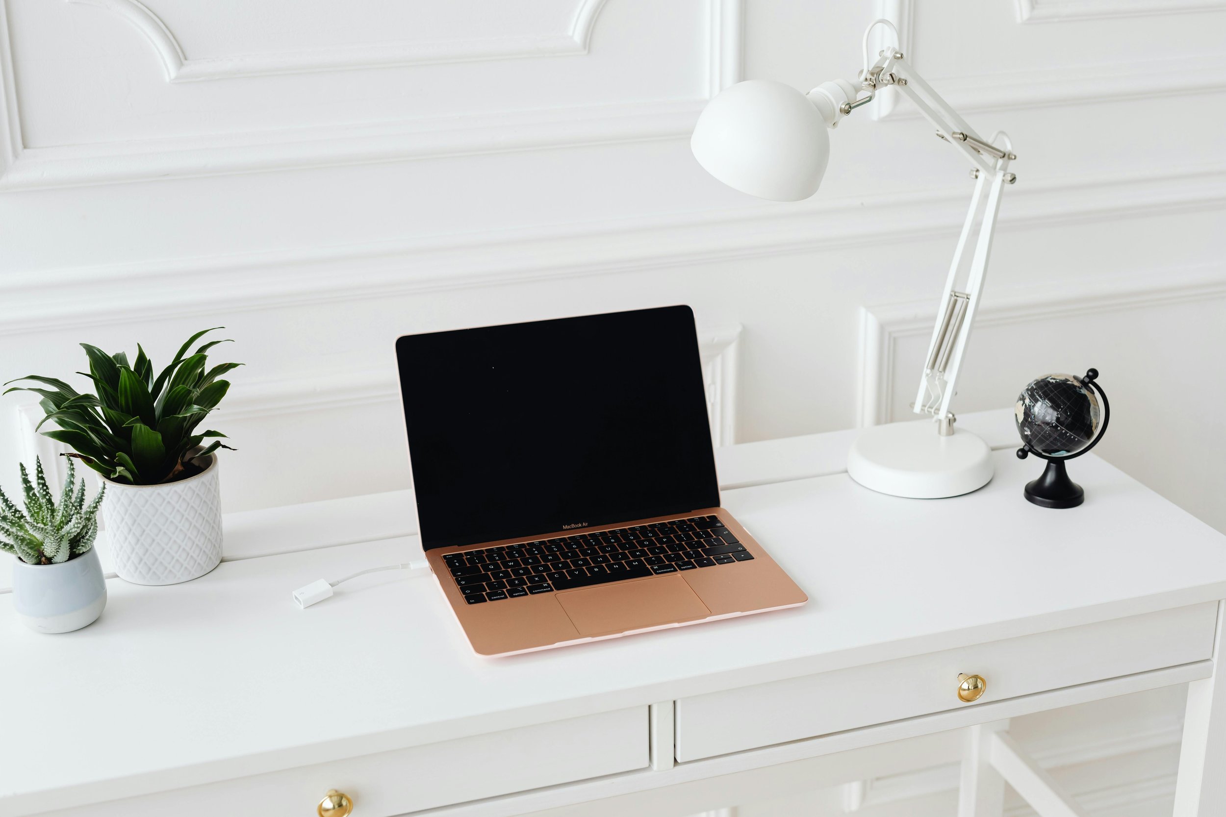 A white desk with a rose gold laptop, two potted plants, a white desk lamp, a small globe, and a drawer with gold knobs, set against a white wall with decorative molding.