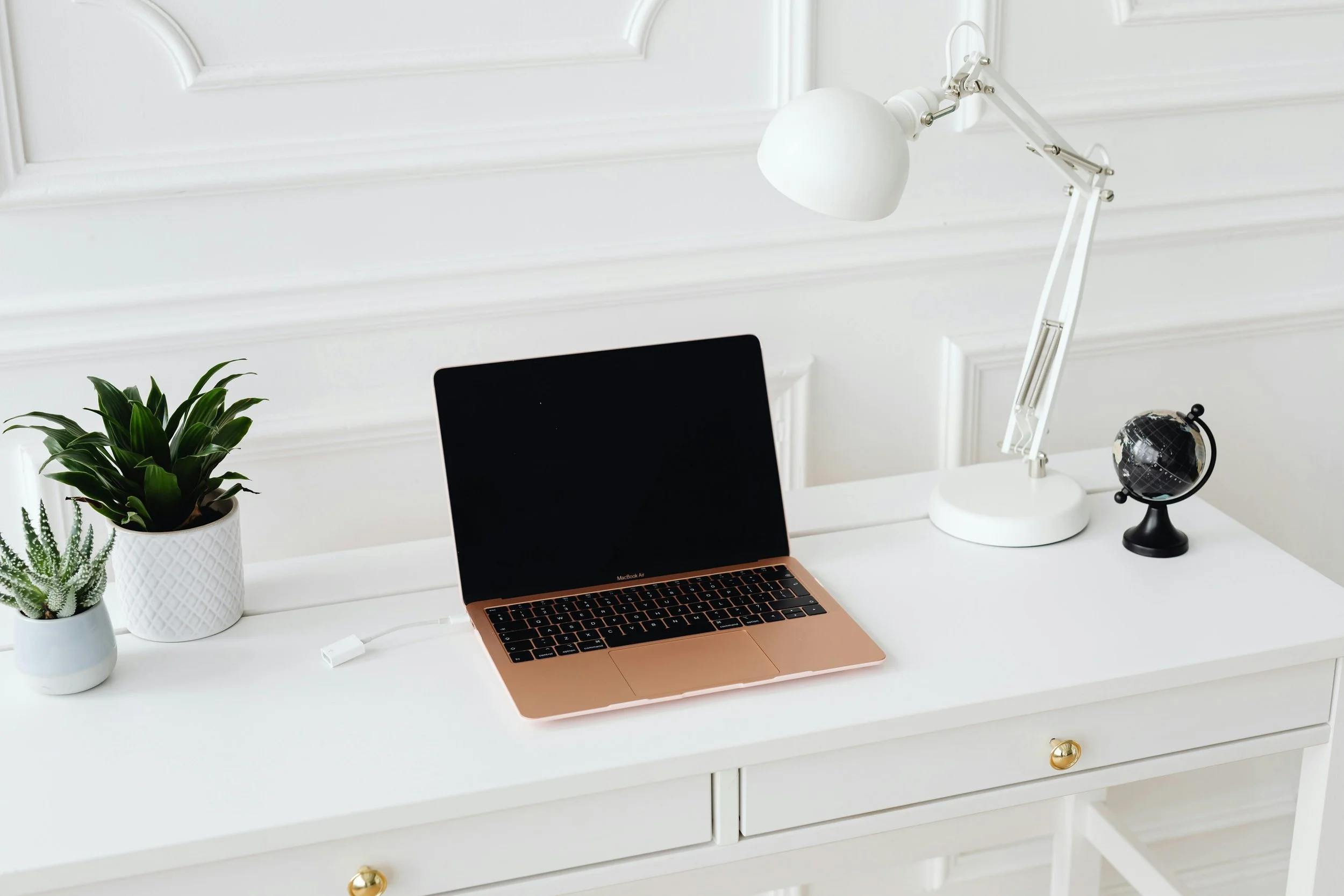 A white desk with a silver laptop, a white desk lamp, a small black globe, and two potted plants in white pots against a white wall with decorative molding.