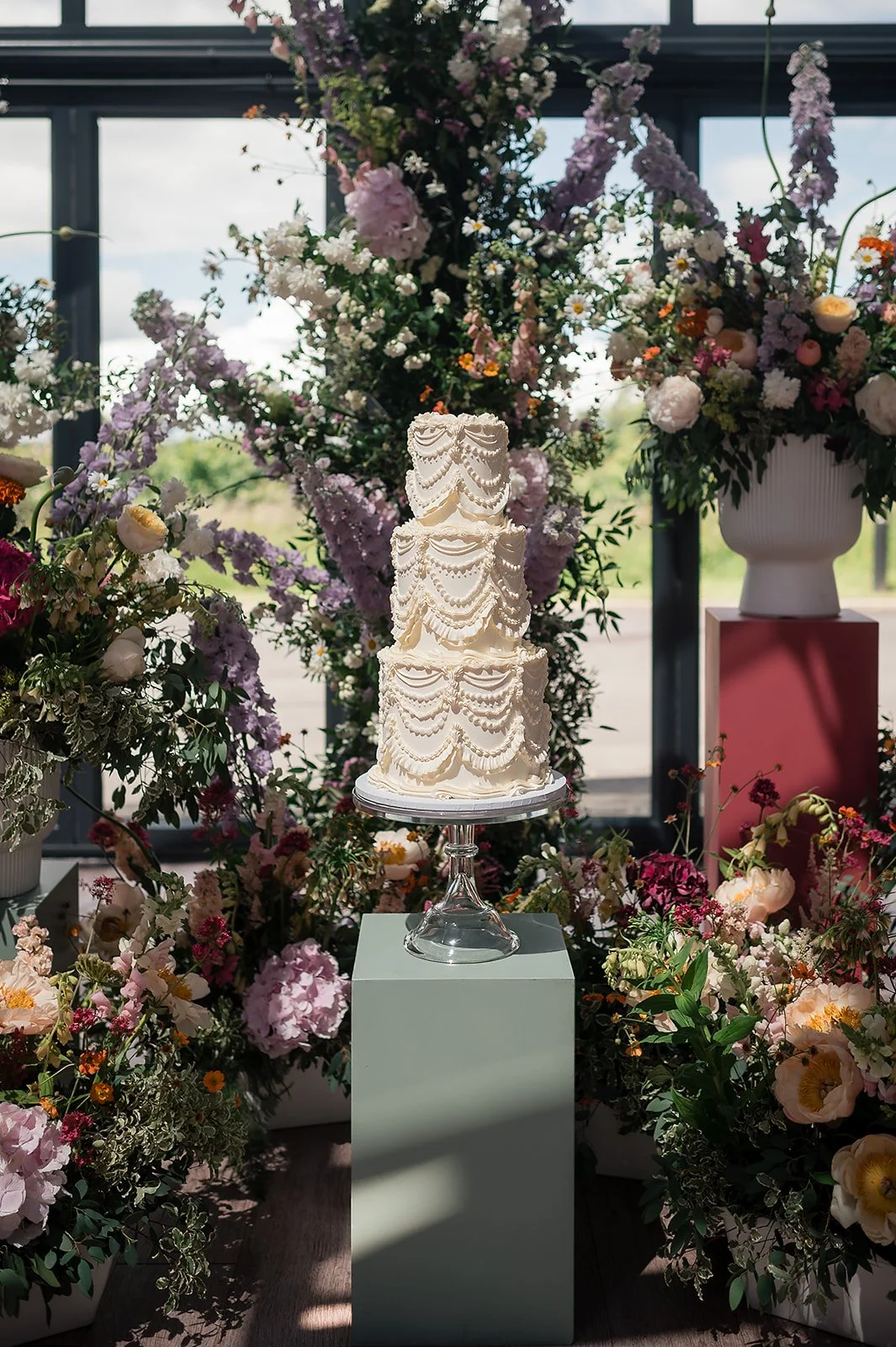 A three-tier white wedding cake decorated with piped icing, placed on a glass cake stand surrounded by an abundance of colorful flowers in vases and arrangements.