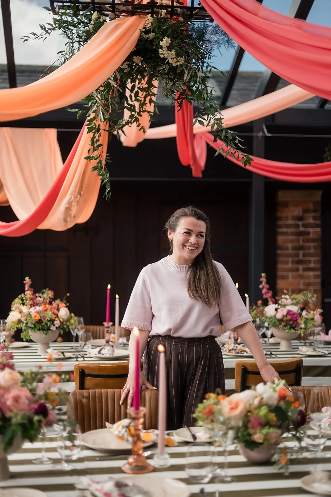 Caroline standing behind a table decorated with floral arrangements and candles at a celebration or party, with pink and peach fabric draped from the ceiling.
