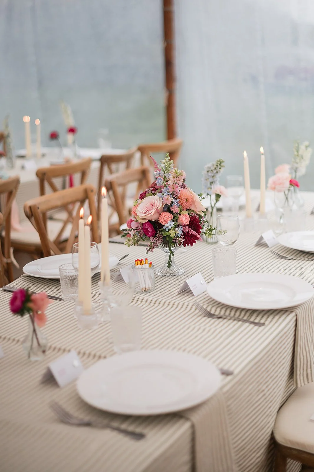 A decorated wedding tablescape with floral centerpieces, white plates, silverware, and candles, set in a bright, elegant marquee.