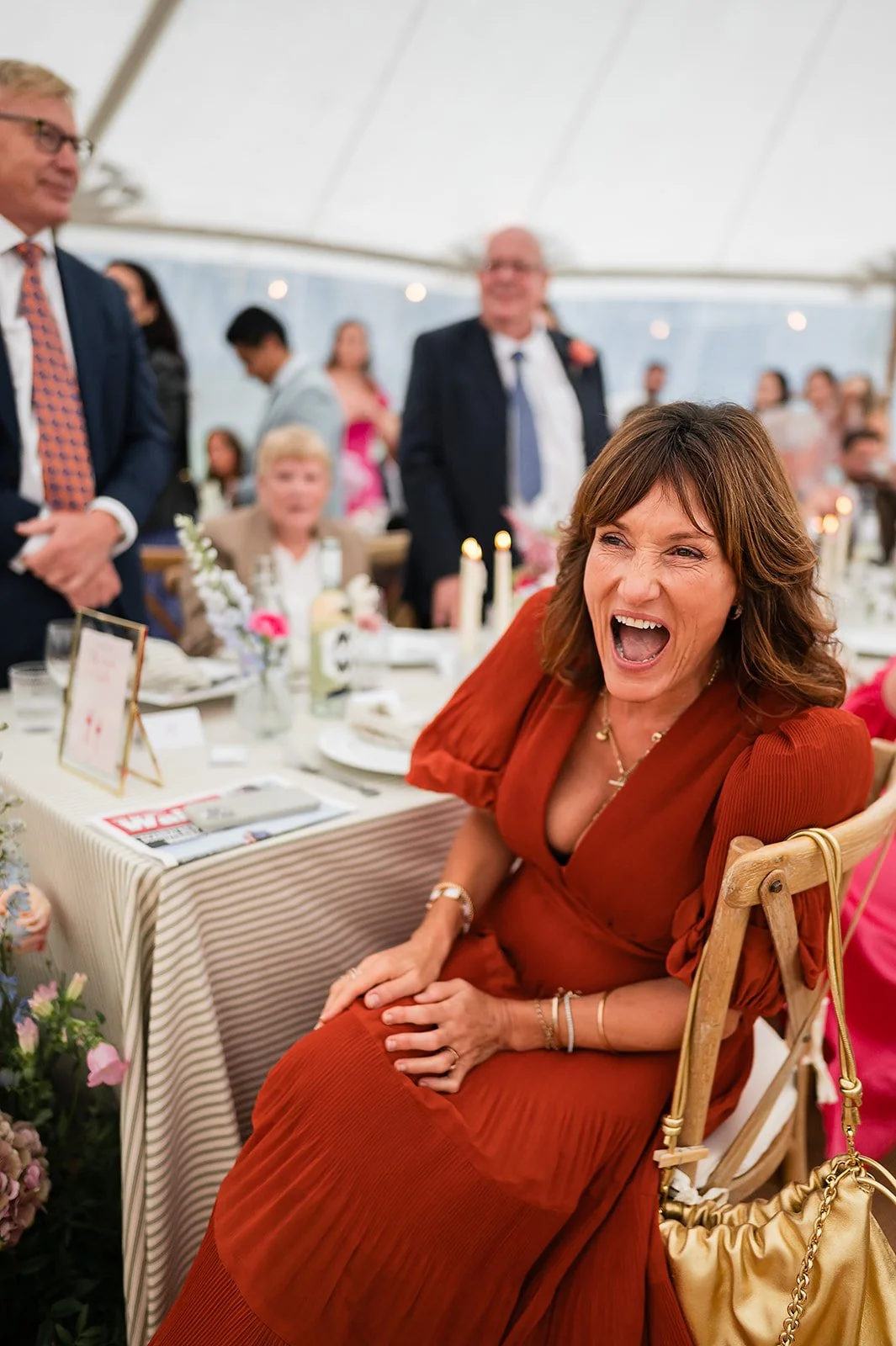 A guest in a rust-colored dress laughing and leaning forward at a table during a celebration or gathering, with several people standing and sitting around her in a marquee setting.
