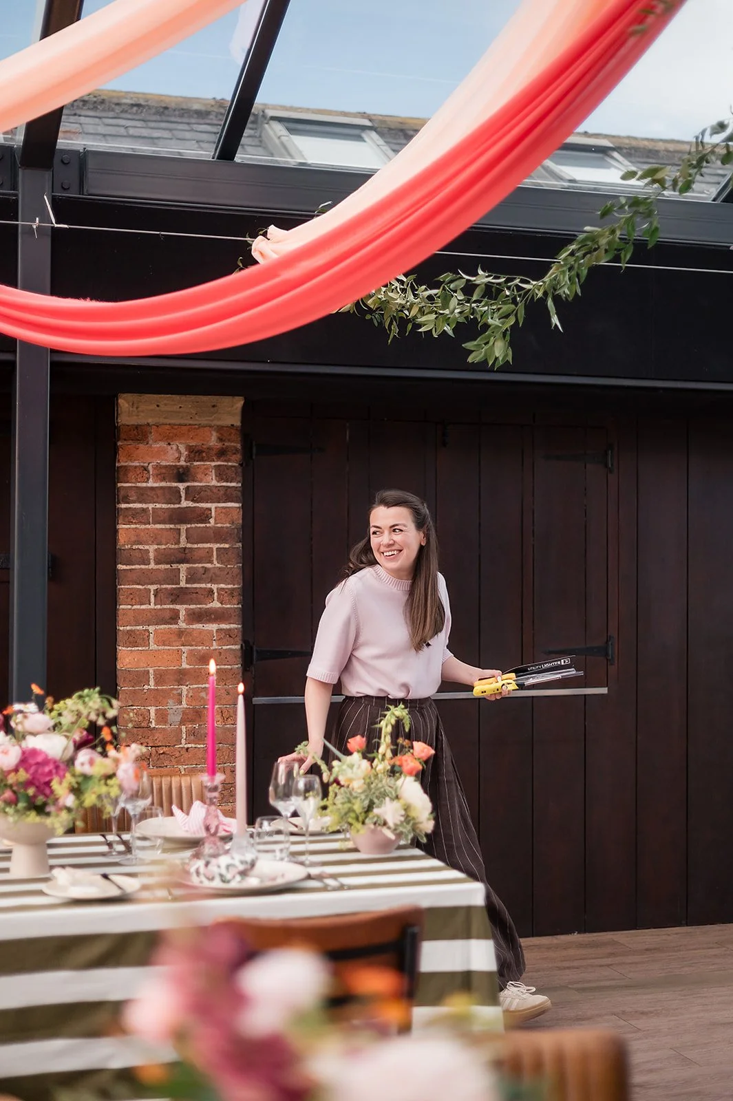 Caroline smiling and holding garden pruning shears, standing near a decorated dining table with floral arrangements and pink candles, in a room with a brick wall and black wooden paneling.