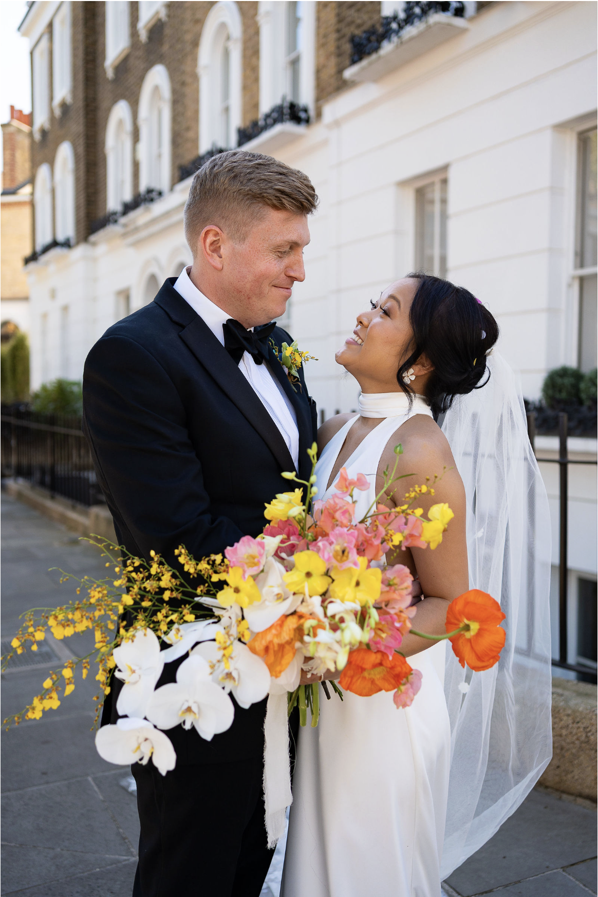 A bride and groom share a moment on their wedding day outside a white building, with the bride holding a large colorful bouquet of flowers.