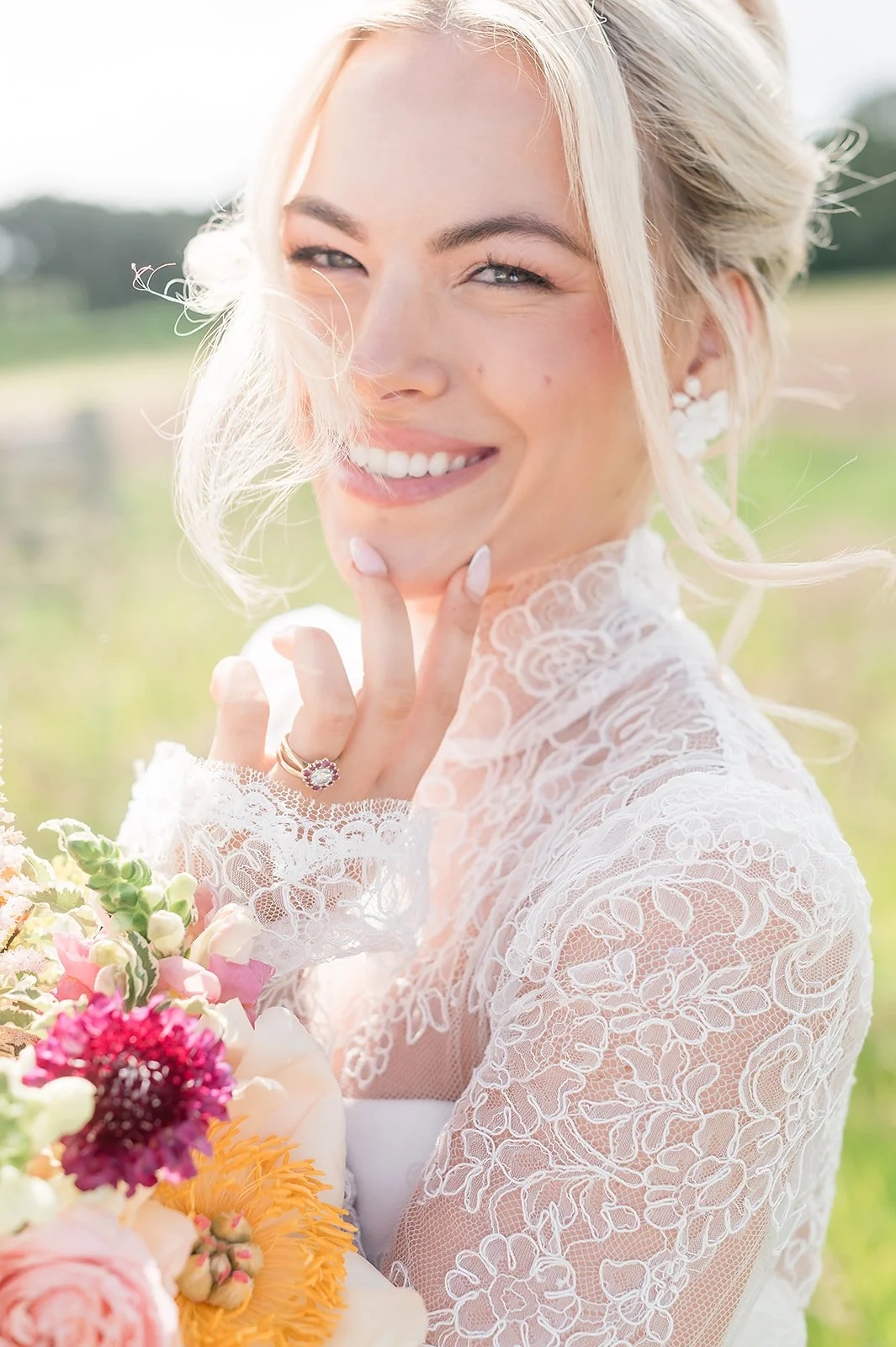 A smiling bride with blonde hair in an outdoor setting, wearing lace clothing and holding a bouquet of flowers.
