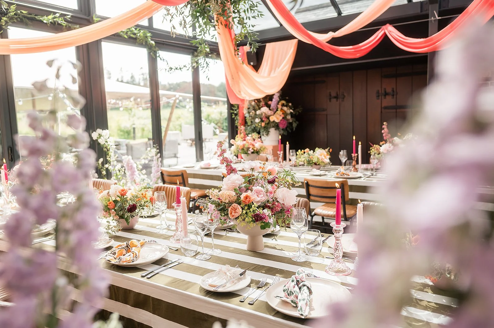A beautifully decorated indoor dining table with pink, peach, and purple floral arrangements, pink candles, and elegant tableware, set in a bright space with large windows and hanging fabric decorations.