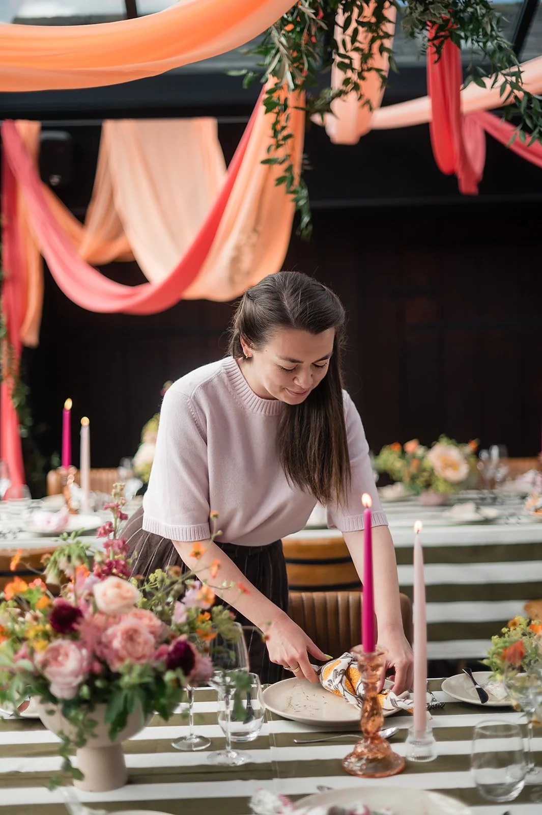 Woman setting a table with pink candles, floral arrangements, and decorated tableware in a decorated venue.