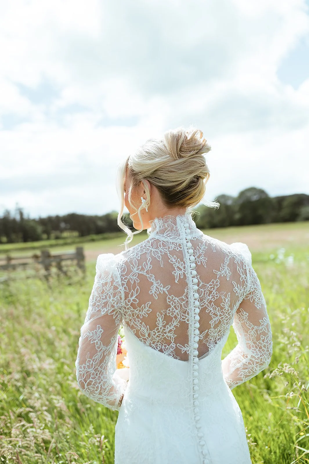 Back view of a bride in a lace wedding dress standing in a field with trees and a cloudy sky.