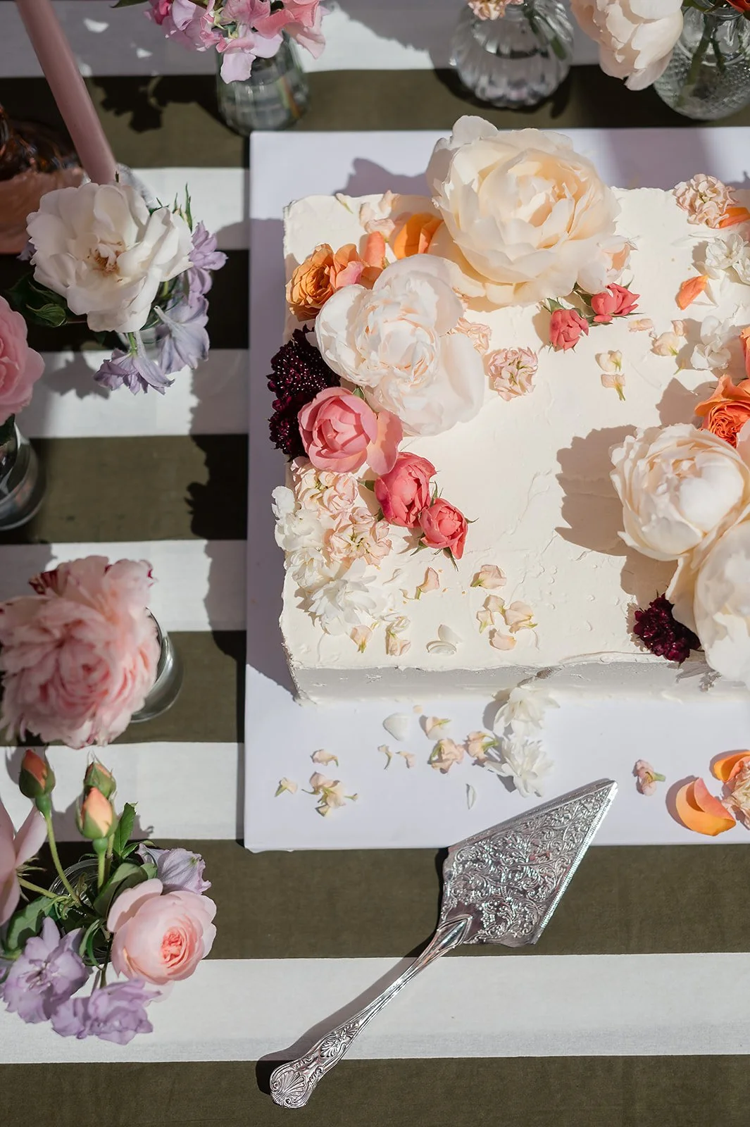A rectangular white cake decorated with large white and pink peonies and smaller pink, peach, and burgundy flowers, topped with flower petals and placed on a white table with floral arrangements nearby.