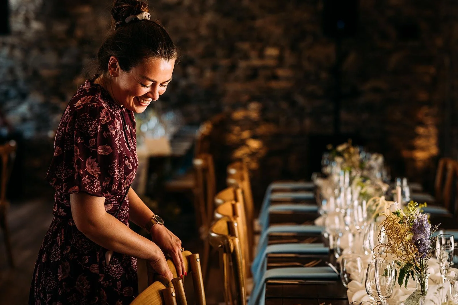 Caroline Judson in a floral dress, setting a long table decorated with floral centerpieces and glassware in a warmly lit rustic indoor setting.