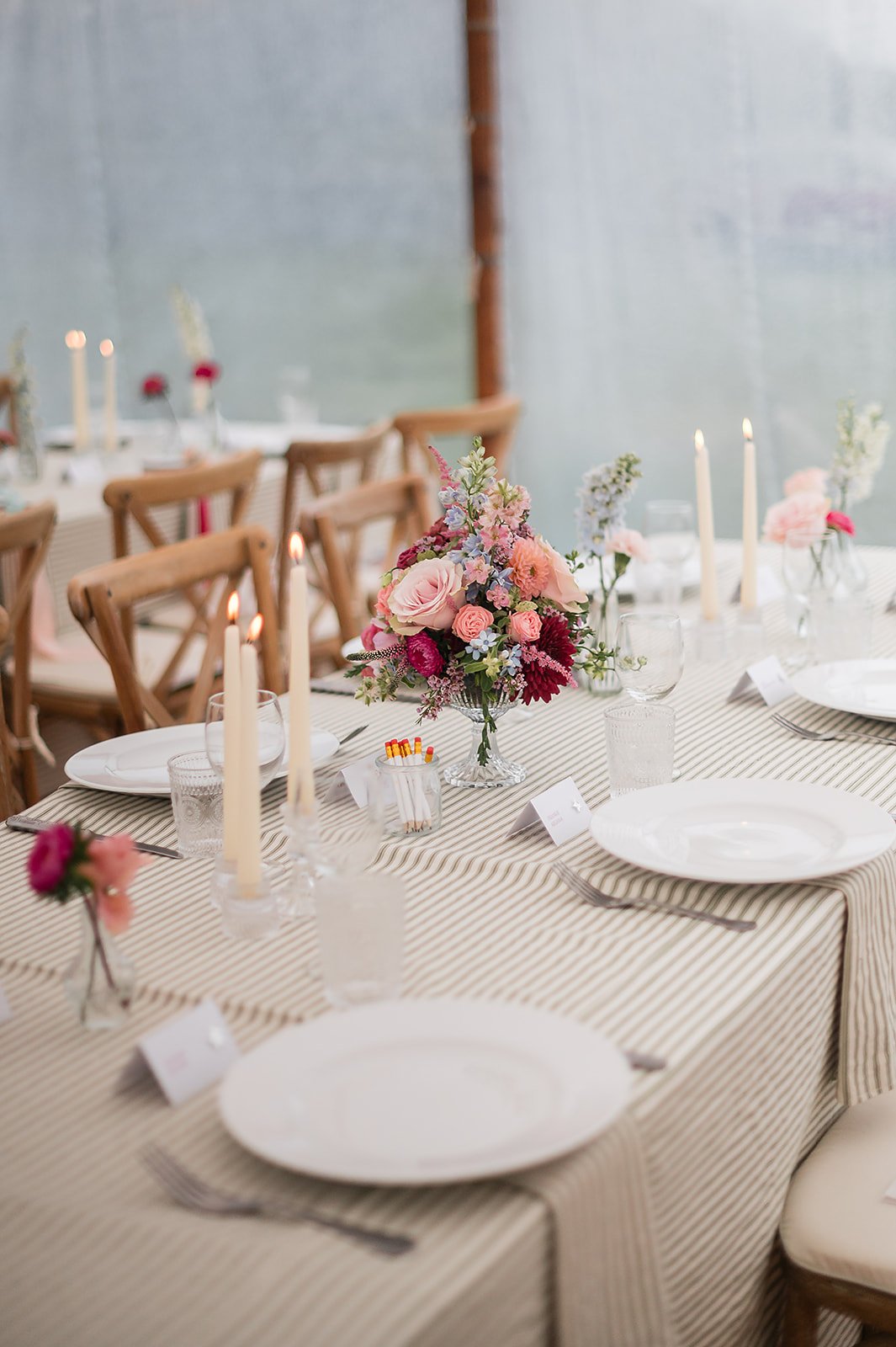 Decorated wedding breakfast table with a floral centerpiece, candles, plates, glasses, and place cards in an outdoor setting.
